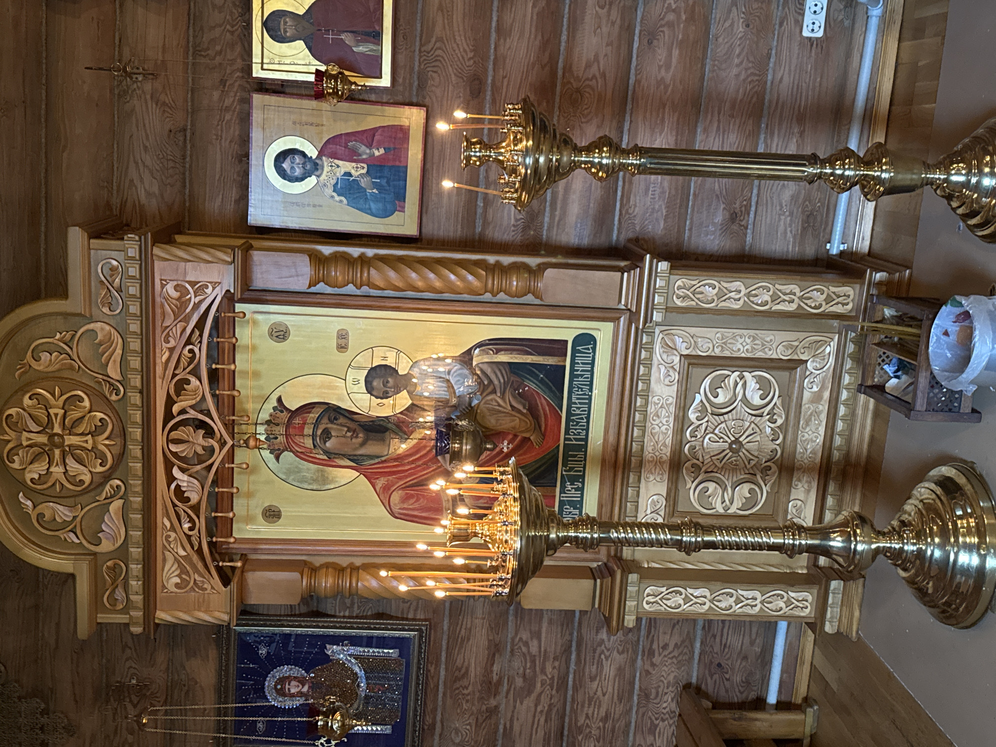 The image shows the interior of a wooden church in Tashla, Russia, likely the Church of the Blessed Mother of God. A large, ornate iconostasis dominates the center of the frame, featuring a central icon of the Theotokos (Virgin Mary) with the Christ Child. Flanking this central icon are smaller icons of saints. Two tall, brass candelabra with numerous lit candles are positioned in front of the iconostasis, one on the left and one on the right. The walls and ceiling are constructed of dark, polished wood. The floor is covered with a light-colored material, possibly a mat or carpet. The overall atmosphere is one of solemnity and reverence, with the warm glow of the candles contributing to the sacred ambiance.