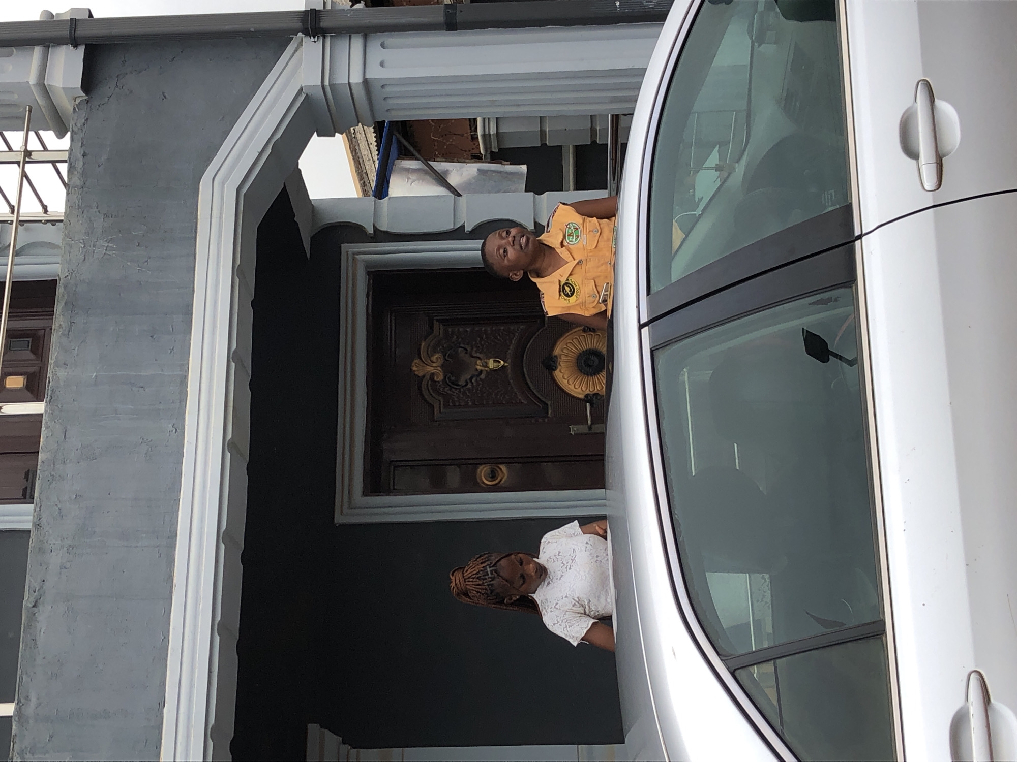 The image captures an outdoor scene featuring two women near a building and a silver car during the daytime. The building has grey walls and ornate white architectural detailing, including a large archway and columns. Behind the archway, a dark wooden door with intricate gold-colored designs and hardware is visible.
On the left, a woman wearing a yellow short-sleeved uniform shirt is seen looking upwards and smiling. A circular patch on her left chest features a green logo with the word "LAGOS" clearly visible, and possibly "SCOUTS" below it. She stands under the archway, seemingly leaning out.
Partially obscured by the car on the right, another woman is visible, wearing a white lace-patterned top and sporting braided hair. She is looking forward, her expression less clear.
A silver car, likely a sedan, occupies the right side of the frame, with its front passenger door, handle, and tinted window clearly visible. The car's roofline and a part of the windshield are also in view. A dark grey gutter pipe runs along the top of the building structure. Some scaffolding or construction elements can be seen in the upper left background. The bright, clear lighting indicates it is daytime.