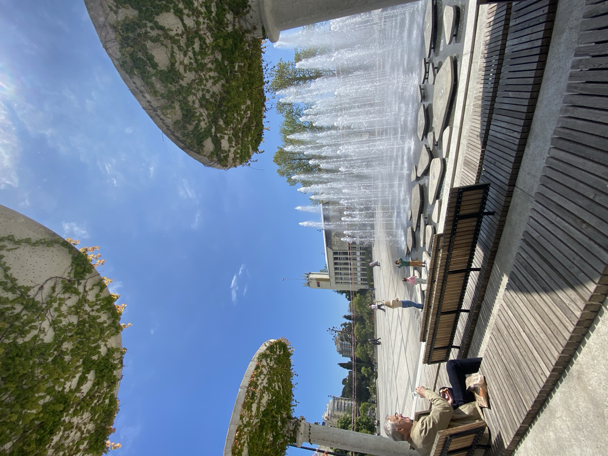 The image depicts a fountain area in город Ялта, Ukraine. Three large, concrete, bowl-shaped structures covered in green vines are arranged in the foreground, framing a view of the sky and the fountain. The fountain consists of numerous jets of water shooting upwards. Behind the fountain is a wide, paved plaza with several people walking. A light-colored, multi-story building is visible in the background. A wooden bench runs along the right side of the frame. An elderly man is sitting on the bench, holding a small object in his hand and facing the fountain.