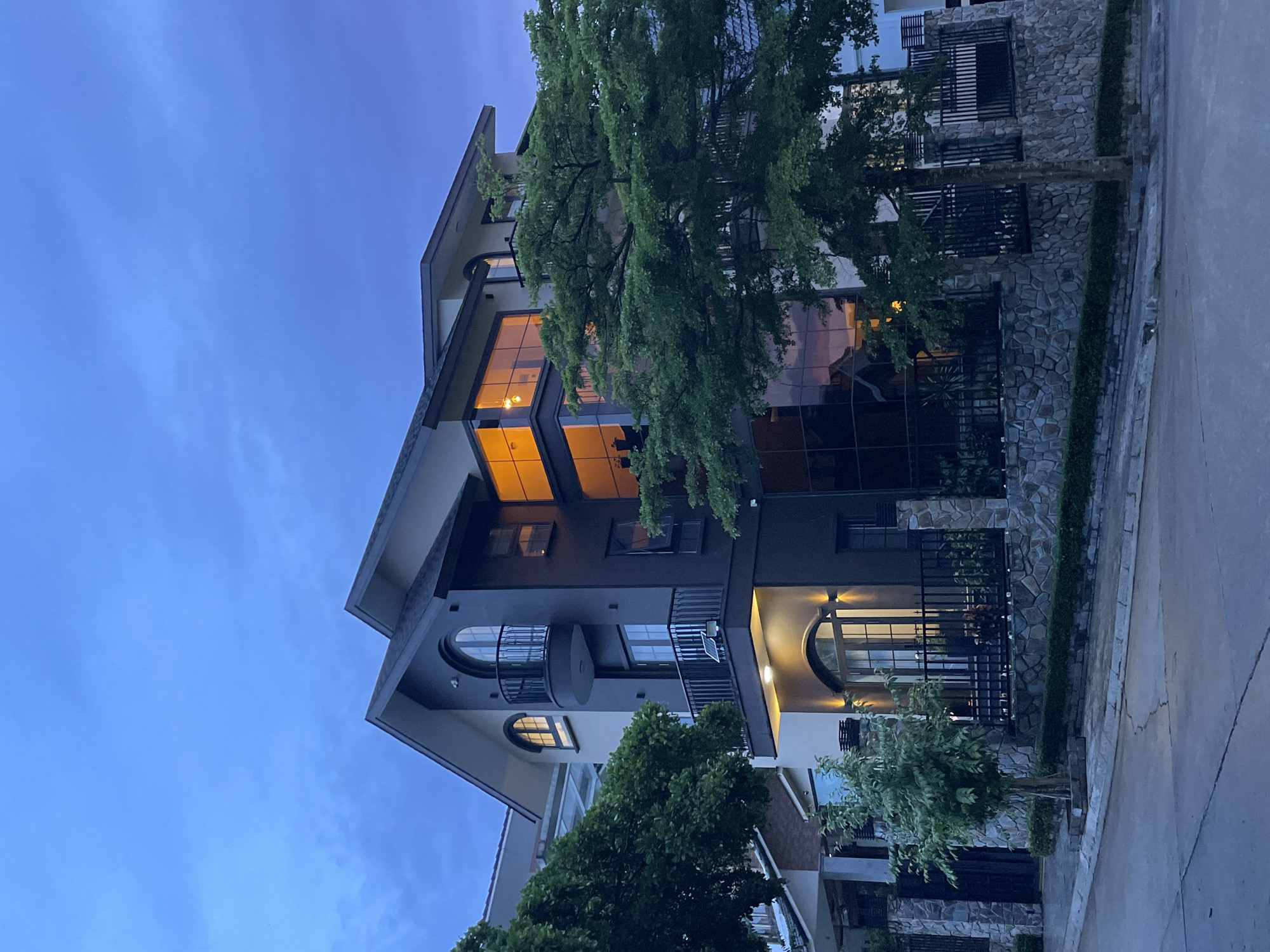 A modern, multi-story house is depicted at twilight on a paved street in the city of Da Lat, Vietnam. The house features dark gray and beige exteriors, large glass windows reflecting the evening sky, and multiple balconies with black railings. A tall tree with lush green foliage stands in front of the house, partially obscuring its facade. The property is bordered by a stone wall and black metal fencing. The sky is a deep blue with wispy clouds, indicating the transition from day to night. There are no people visible in the image. The environment appears to be a residential area with other houses visible in the background.