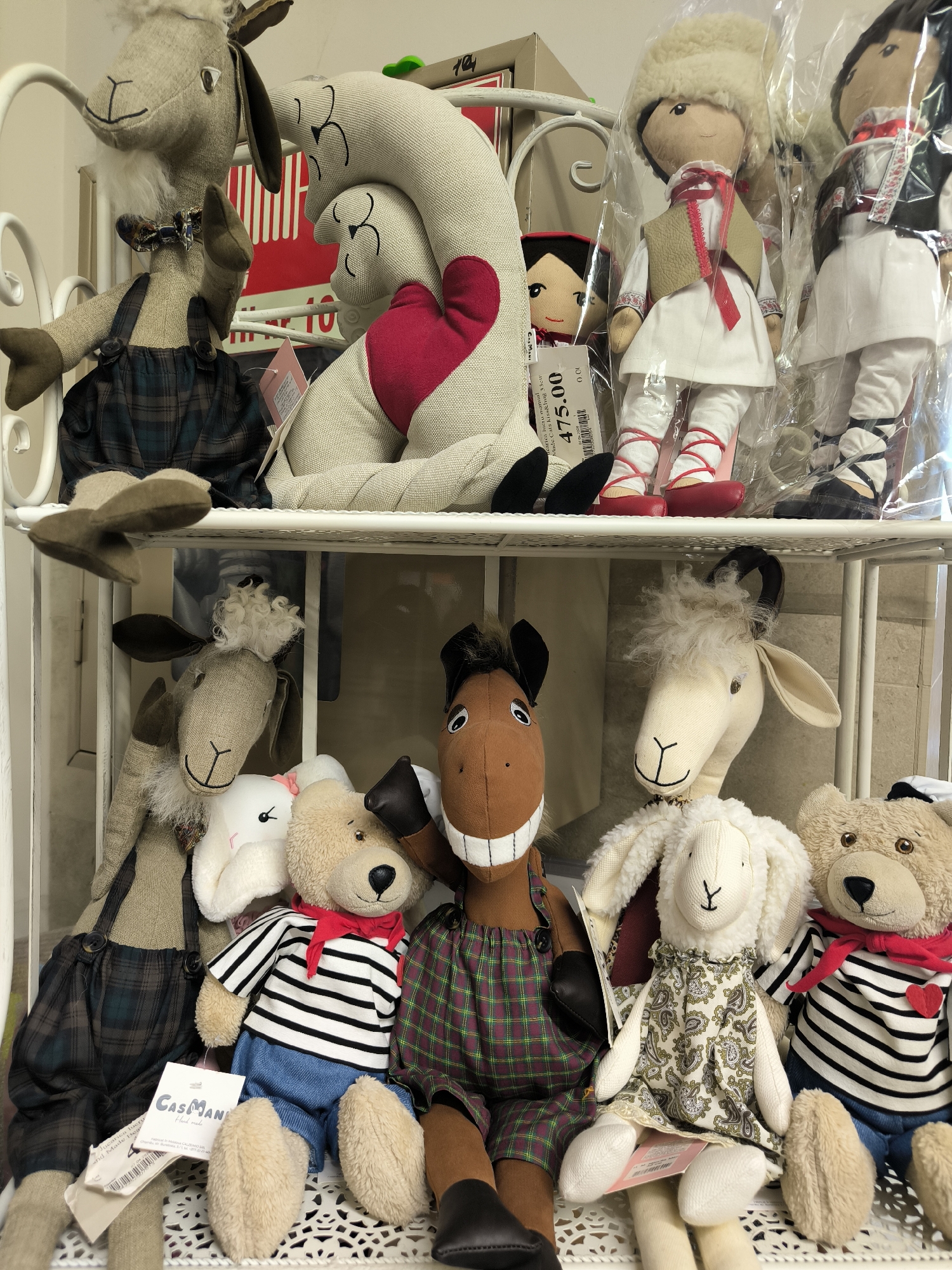 The image shows a collection of plush toys arranged on white metal shelves. On the top shelf, a large stuffed sheep with a red heart is prominent, alongside two dolls in traditional attire. Below, the bottom shelf features a stuffed horse, several sheep, and two teddy bears. A tag that reads "CasaMan" and "Hand made" is visible on one of the sheep. The setting appears to be a shop or market stall. There is no visible indication of the specific location within Chișinău, Moldova, beyond it being an indoor retail environment.