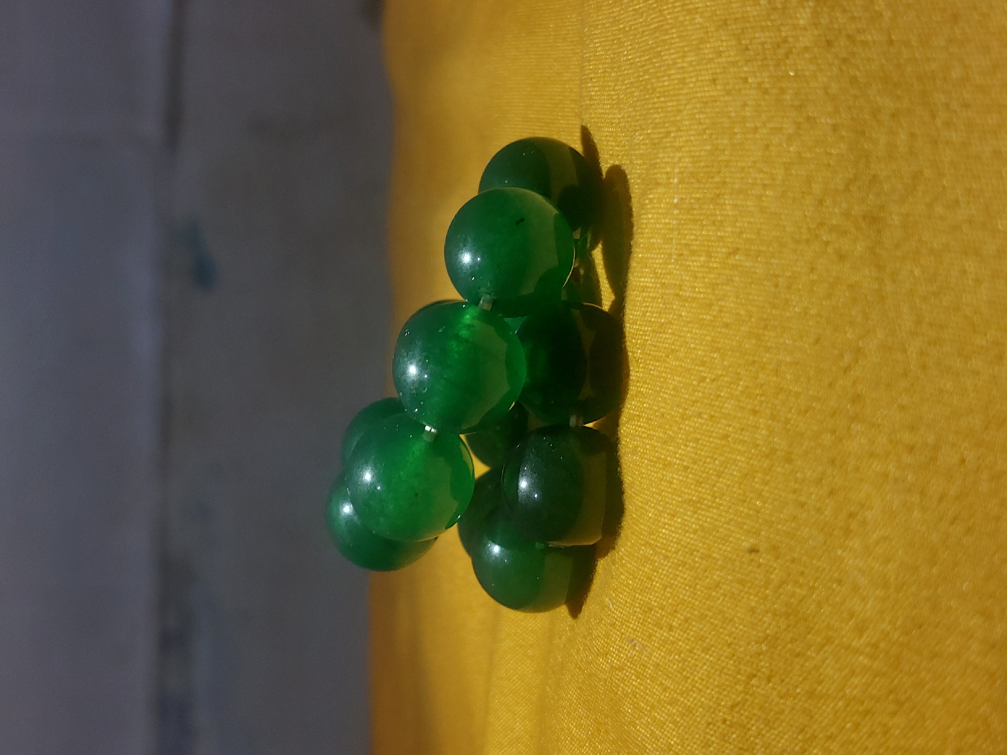 The image shows a stack of approximately nine green, translucent, spherical beads on a textured, yellow surface. A dark shadow is cast to the right of the beads. The background on the left side of the image is a light gray, possibly a wall or other flat surface. The image does not provide any specific information to determine the location in the city of Lhokseumawe, Indonesia, or any subjects, people, or interactions.