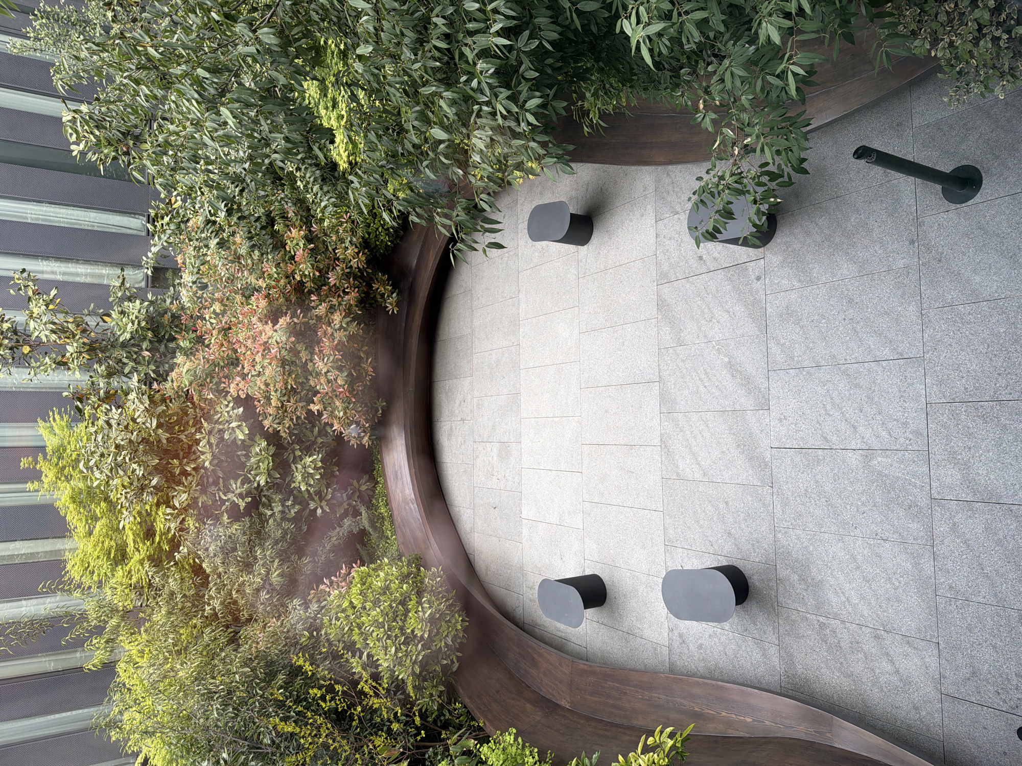 Captured from an elevated perspective, the photograph presents an outdoor urban space in Tbilisi, Georgia. The location features a curved wooden planter filled with diverse greenery including shrubs and trees. The planter borders a tiled gray patio. Four modern black benches are positioned along the patio's edge. In the upper right corner, a black object, possibly a pipe or fixture, is mounted on the tiled surface. In the background, a building with glass panels is visible. Sunlight filters through the foliage, creating a dappled lighting effect on the plants and patio surface. No people are present in the image.