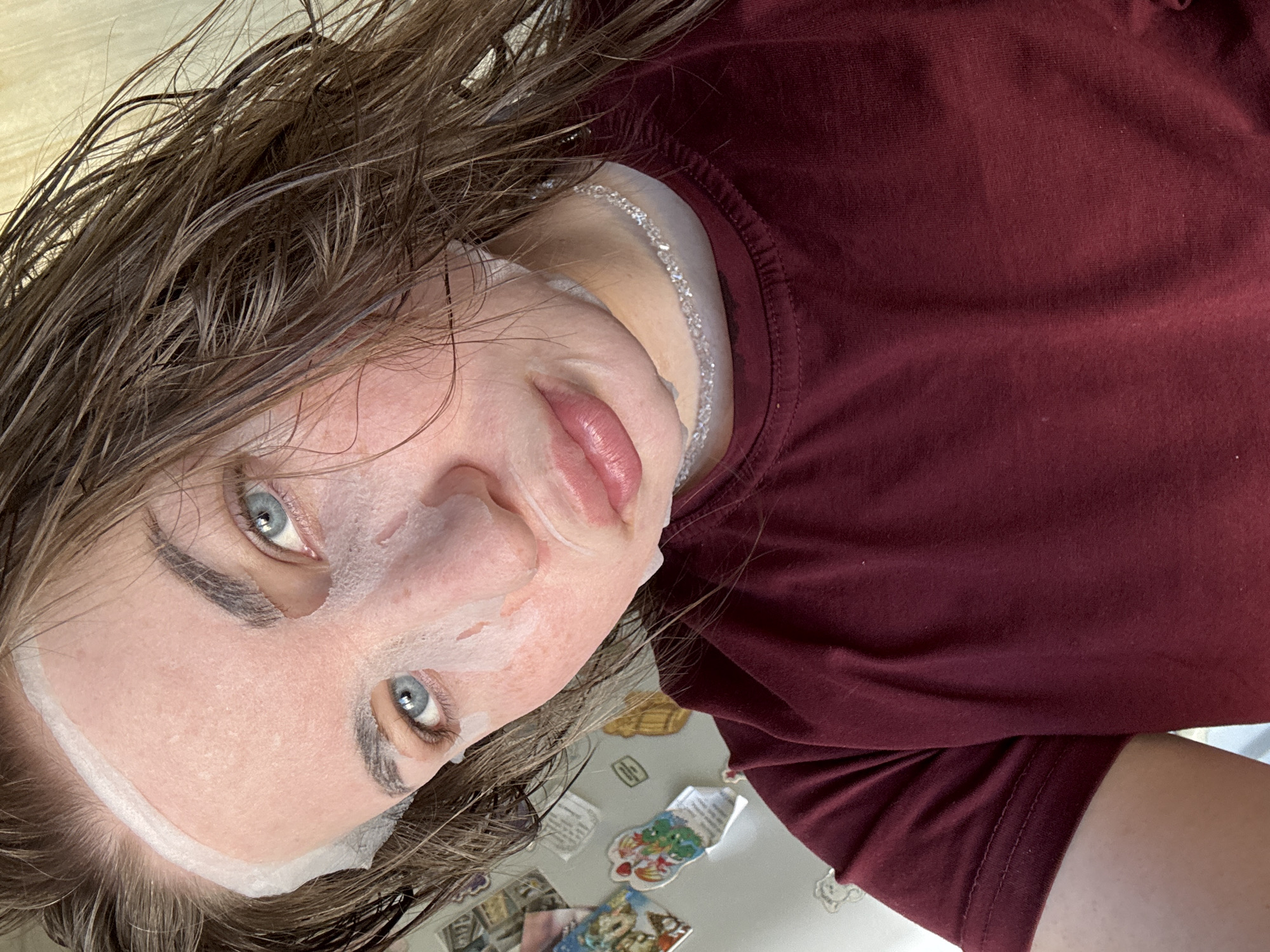 A selfie portrait of a person with wet brown hair wearing a white sheet face mask and a maroon short-sleeved shirt. The person has fair skin, visible eyebrows, and blue eyes, looking directly at the camera. The background is a white surface with several small, colorful magnets.