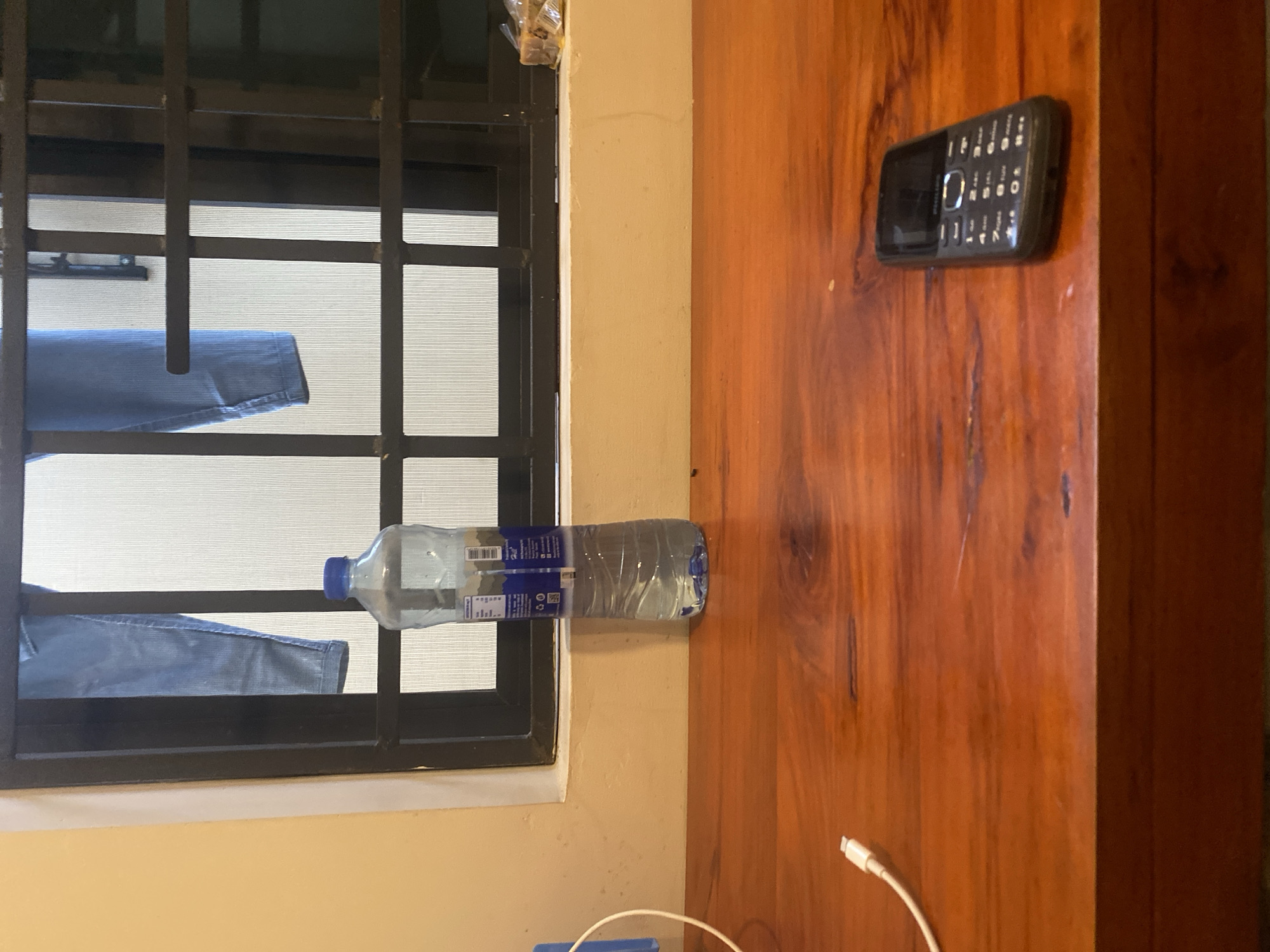 The image captures an indoor scene, primarily divided between a window on the left and a wooden surface on the right. The scene is lit by diffused daylight coming through the window, suggesting it's daytime.
On the left, a window with black metal security bars is prominent. Through the bars, a light-colored, possibly screened or blinded surface is visible, along with what appears to be a pair of blue jeans or trousers draped over one of the lower horizontal bars, airing or drying. Above the window frame, a small, crumpled, yellowish item in clear plastic is tucked into the corner.
Resting horizontally across the bottom section of the window frame and the adjacent beige wall is a clear plastic water bottle with a blue cap. The bottle, which appears mostly full, has a label with blue text and graphics, partially revealing "SMARTWATER" and "Electrolytes for taste", along with other unreadable nutritional information and a barcode.
To the right, a reddish-brown wooden surface, likely a cabinet or door, dominates the space. This surface exhibits distinct wood grain patterns and some minor scratches. Placed near the top right of this wooden surface is a black feature phone, resembling an older Nokia model, with a physical keypad and a small screen. At the bottom of the wooden surface, a white charging cable, possibly an Apple Lightning cable, is visible, curving upwards from the bottom edge of the frame.
No people are present, and the overall scene suggests a functional, domestic setting. The security bars on the window could be a common feature in the location, which is Dar es-Salaam, Tanzania.