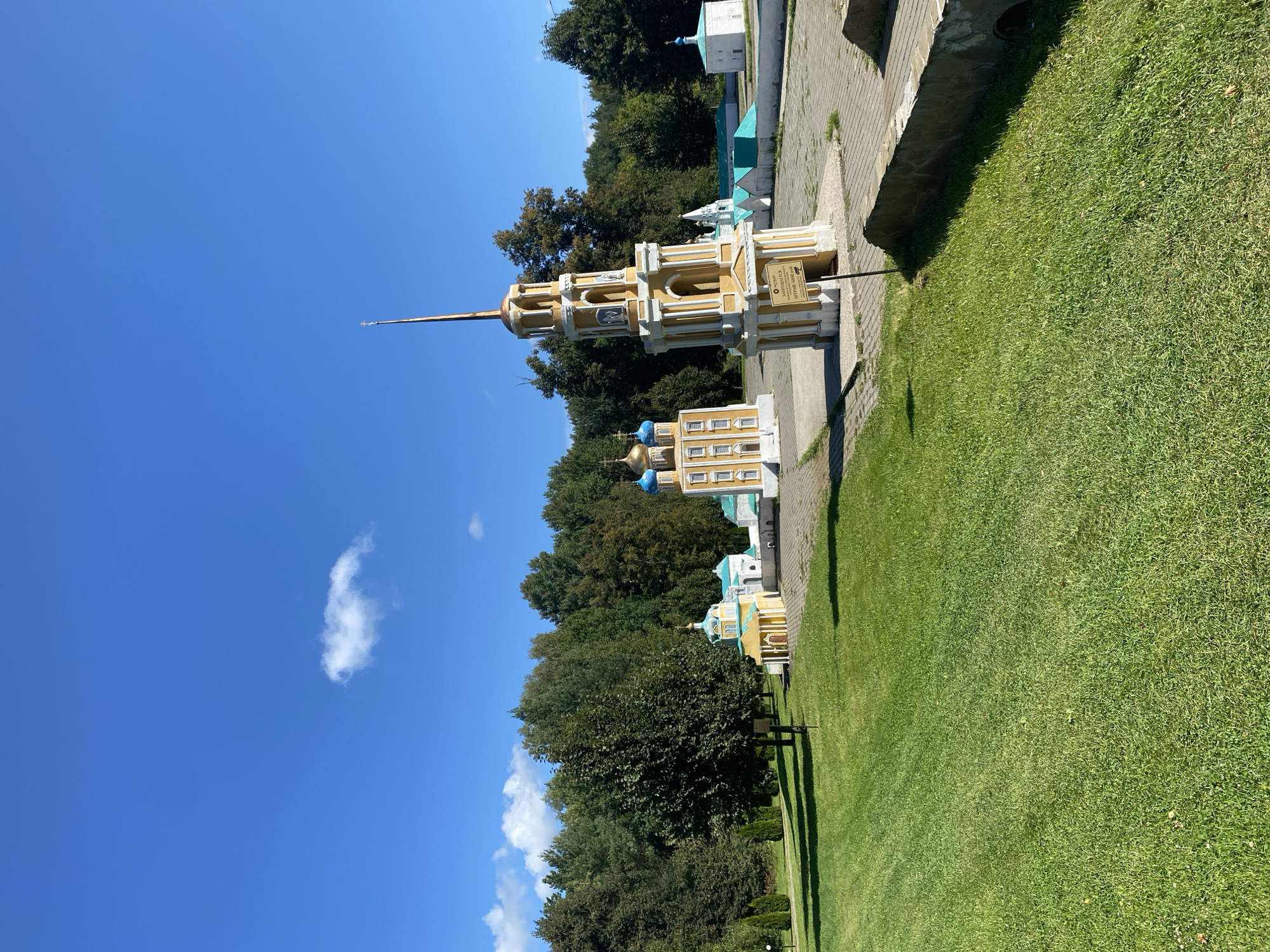 The image depicts an outdoor scene on a sunny day with a clear blue sky and a few scattered clouds. The foreground features a well-maintained green lawn. In the midground and background, there are several miniature architectural models, resembling churches and bell towers. These models are colorful, with yellow and white facades, and some have blue or teal domes. One prominent model is a tall, multi-tiered yellow bell tower with a spire. Behind these models, a dense line of green trees forms a natural backdrop. The ground around the models appears to be paved with grey interlocking stones. There are no people visible in the image. The scene is likely a park or an open-air museum. The location is identified as Chulis, Russia.