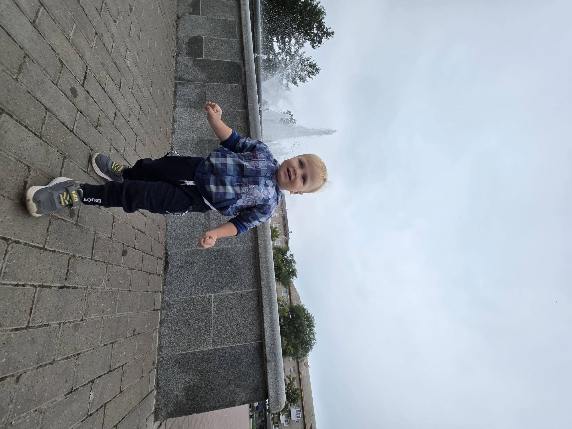 A low-angle medium shot shows a fair-haired toddler boy running towards the camera on a gray brick sidewalk. He is wearing a blue plaid hooded jacket with white trim, dark blue pants with white lettering, and gray sneakers with yellow laces. Behind him, a large, ornate fountain sprays water into the air. The fountain is bordered by a gray stone wall, beyond which are trees and buildings. The sky above is overcast and pale gray. The image appears to have been taken in a public park or plaza. The boy is smiling and appears to be enjoying himself. The background suggests an urban setting, potentially in Chaikovsky, Russia, given the presence of typical urban architecture and the style of the fountain.