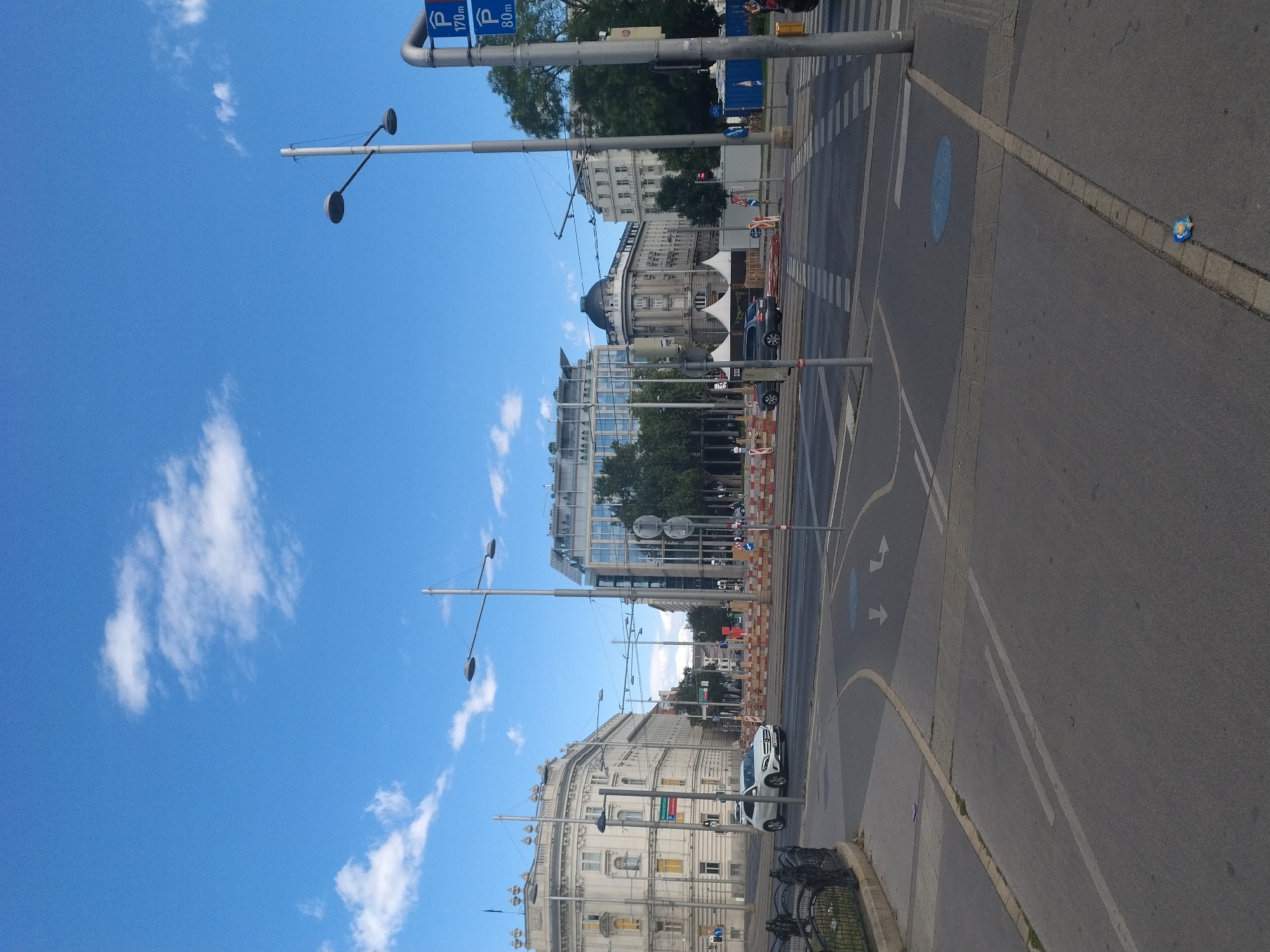 The image shows a street scene in Vienna, Austria, likely near a major intersection or public square, given the presence of multiple lanes, crosswalks, and traffic signals. In the foreground, a grey asphalt road is visible, with white lane markings and pedestrian crossings. A white car is driving on the road, and to the left, there is a black metal fence bordering a grassy area.
The background features several multi-story buildings. On the left, a light-colored building with ornate architecture stands prominently. In the center, a modern building with a glass facade and a dark-domed building are visible. The sky is blue with scattered white clouds.
There are several streetlights and utility poles throughout the scene. Traffic construction barriers, consisting of orange and white striped elements, are present, suggesting ongoing work in the area. A few pedestrians are visible in the distance, walking near the buildings. Two blue parking signs with white "P" symbols and distances (170m and 80m) are mounted on a pole on the right side of the image. A discarded wrapper is visible on the ground in the lower right portion of the frame.