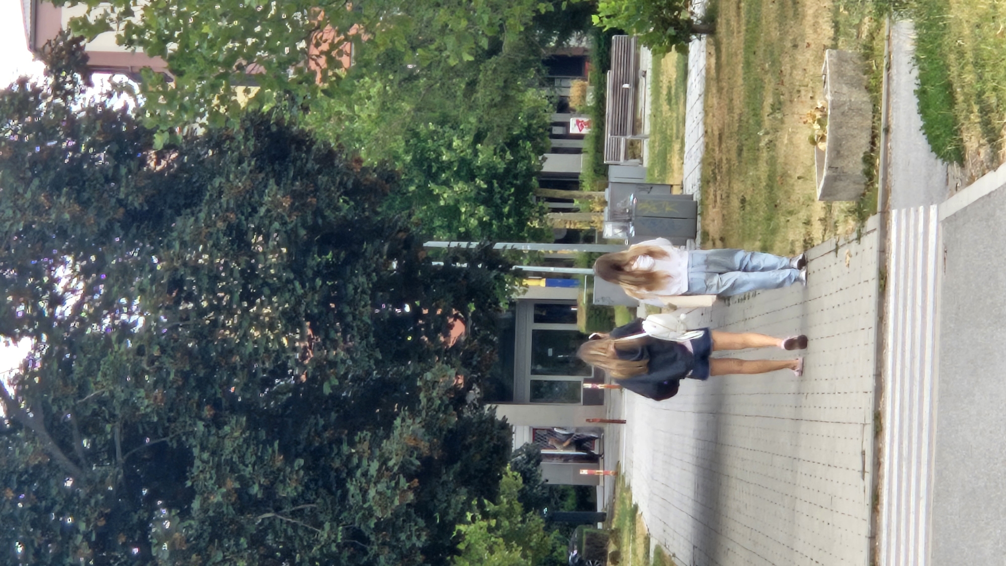 The image captures a street scene with two young women walking away from the camera on a paved walkway. The woman on the left wears a dark hoodie, denim shorts, and pink flip-flops, carrying a white backpack. The woman on the right is dressed in a white hoodie, light blue jeans, and white sneakers, also carrying a light-colored bag. They are walking towards a building with large glass windows and a red door visible in the background, partially obscured by trees. A pedestrian crossing is visible in the foreground. The environment includes green trees, grass, and urban elements like benches and trash receptacles. The scene appears to be in an outdoor, public space, possibly near a school or a university campus, given the presence of students. The image does not provide specific visual cues to definitively place it in Prešov, Slovakia.