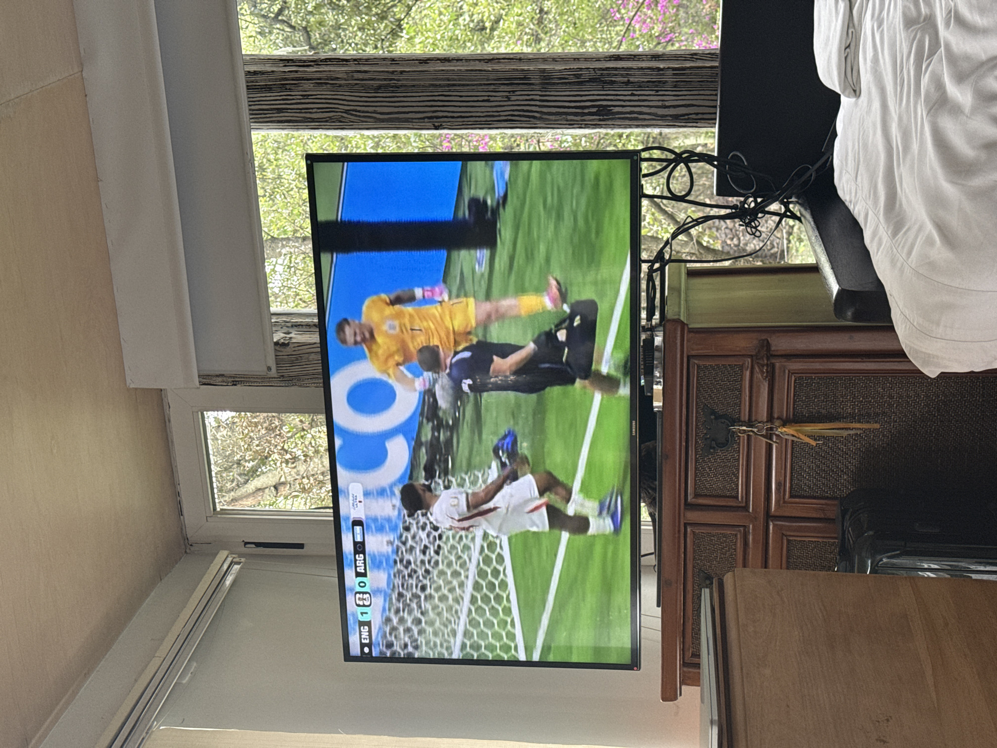 An indoor scene depicts a television screen displaying a soccer match. The screen shows a score overlay indicating "ENG 1 0 ARG". Two male figures are visible on the screen: one in a yellow goalkeeper uniform and another in a dark blue kit. The television is placed on a wooden dresser with ornate carvings. To the left of the dresser, a wooden cabinet is partially visible, and to the right, a black headboard of a bed is in view with white bedding. Through a window behind the television, lush green trees and some pink flowering branches are visible, suggesting an outdoor environment with abundant vegetation. The overall lighting suggests it is daytime. The image does not contain any information that would allow for the determination of its location in Mexico City, Mexico.