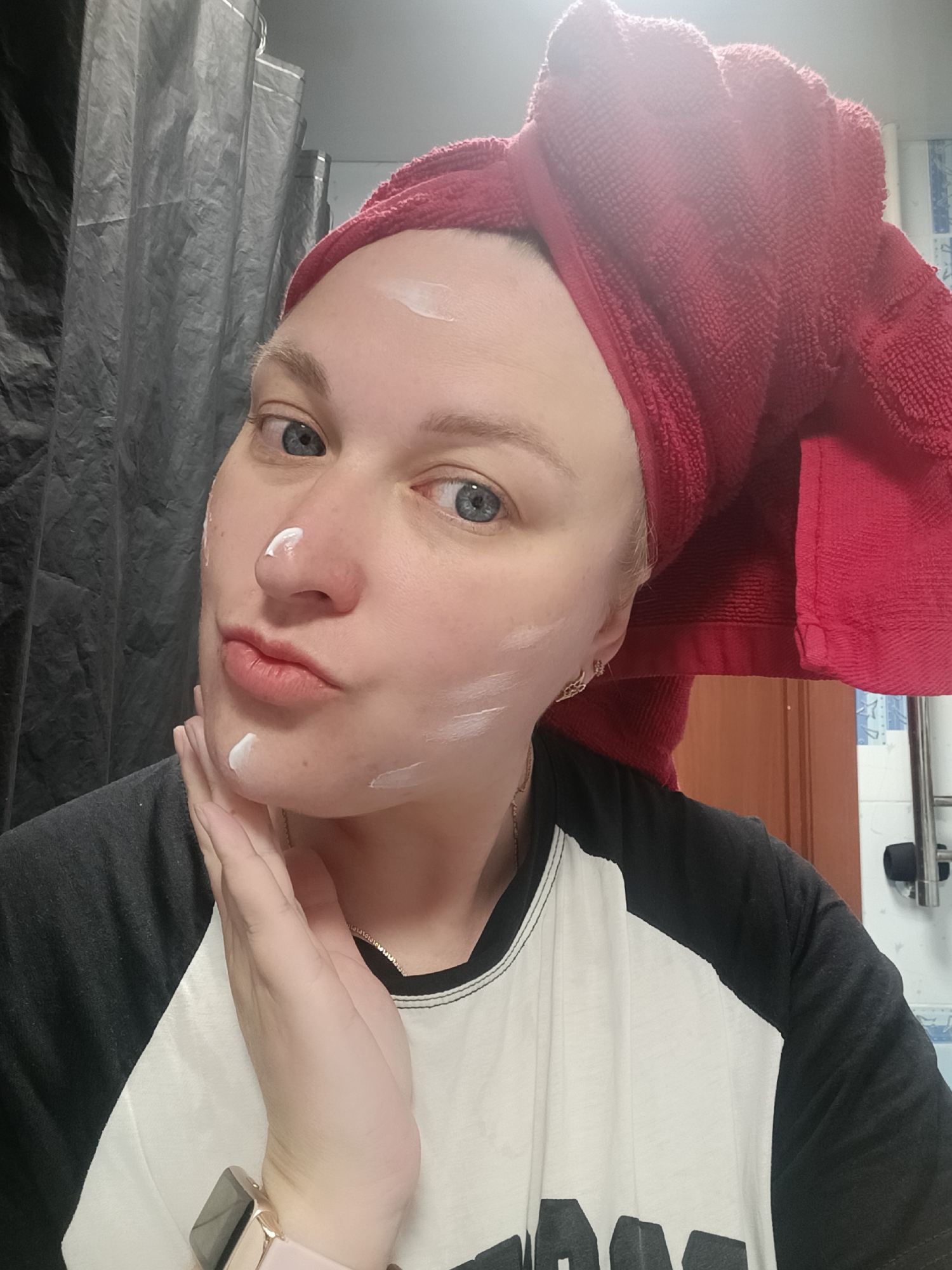 A woman with fair skin, blue eyes, and blonde hair wrapped in a red towel is taking a selfie. She has white cream or lotion applied on her forehead, nose, and cheek. She is wearing a black and white baseball tee. Her right hand is touching her chin, and she is pouting her lips. A gold necklace is visible on her neck, and a smartwatch with a pink band is on her left wrist. In the background, there is a gray shower curtain, a wooden door, and a towel rack with blue and white tiles on the wall. The location is likely a bathroom. The city of Chaikovsky, Russia, cannot be verified from the image.