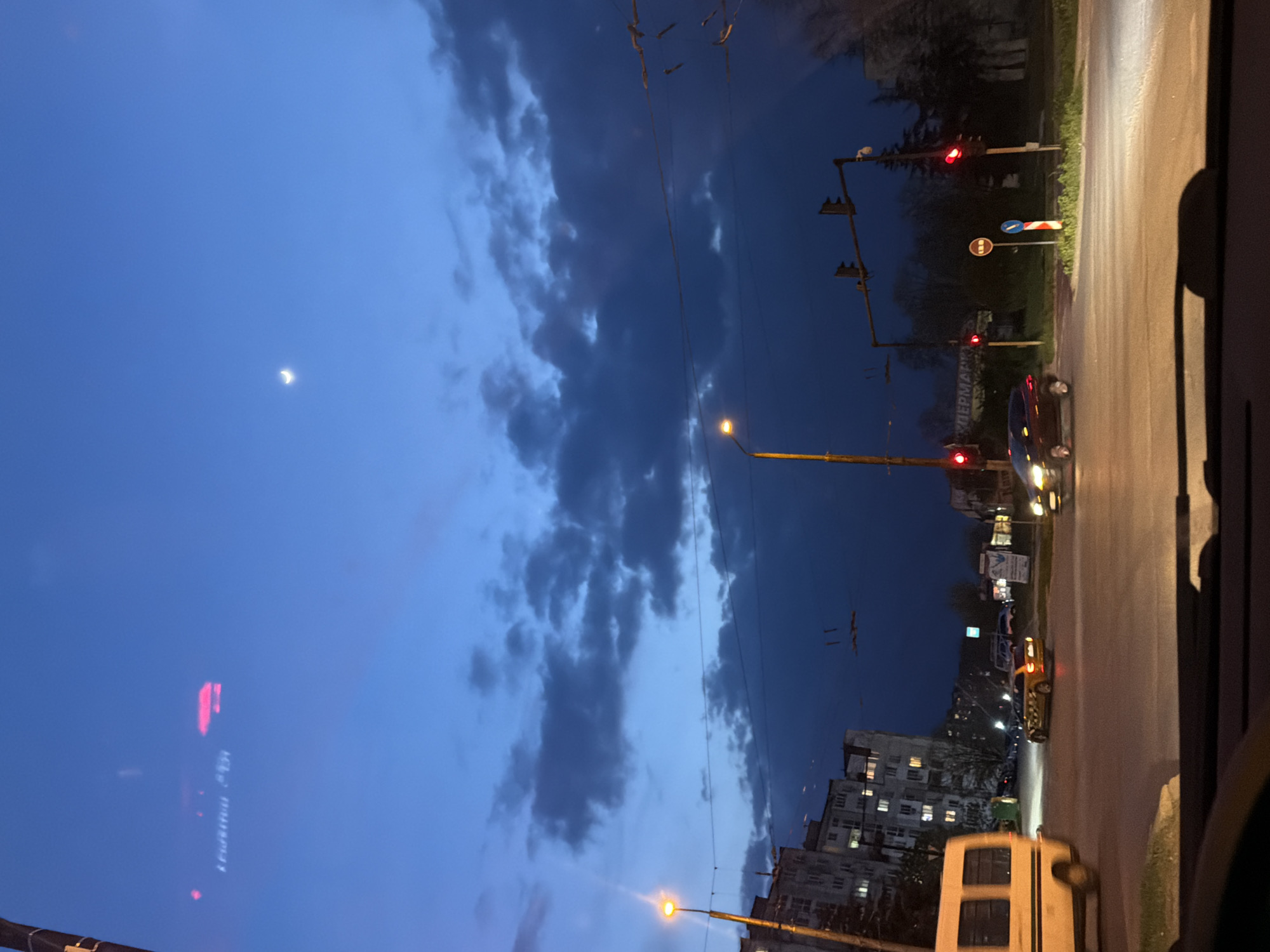 The image captures an urban street scene at dusk or early night, viewed from inside a vehicle. The sky transitions from a lighter blue, possibly towards the west, to a darker, deep blue overhead, with scattered dark clouds. A distinct crescent moon is visible in the upper part of the sky.
On the ground level, a street runs across the frame, illuminated by the warm glow of streetlights and vehicle headlights. Several vehicles are present, including a dark car stopped at a red traffic light, a yellow taxi further down the street, and the partial view of a white van-like vehicle in the foreground. Multiple utility poles with power lines crisscross the upper part of the scene, supporting traffic lights that are currently red. Various road signs are visible, including a red circular 'no entry' or 'stop' sign, and a blue circular sign with a white arrow.
In the background, multi-story residential buildings with illuminated windows suggest an active urban environment. Cyrillic text is clearly visible on a sign near the road, spelling "СУПЕРМАРКЕТ" (Supermarket). This, combined with the location context provided, confirms the setting as Varna, Bulgaria. The overall atmosphere is quiet and observant, capturing a typical city evening.