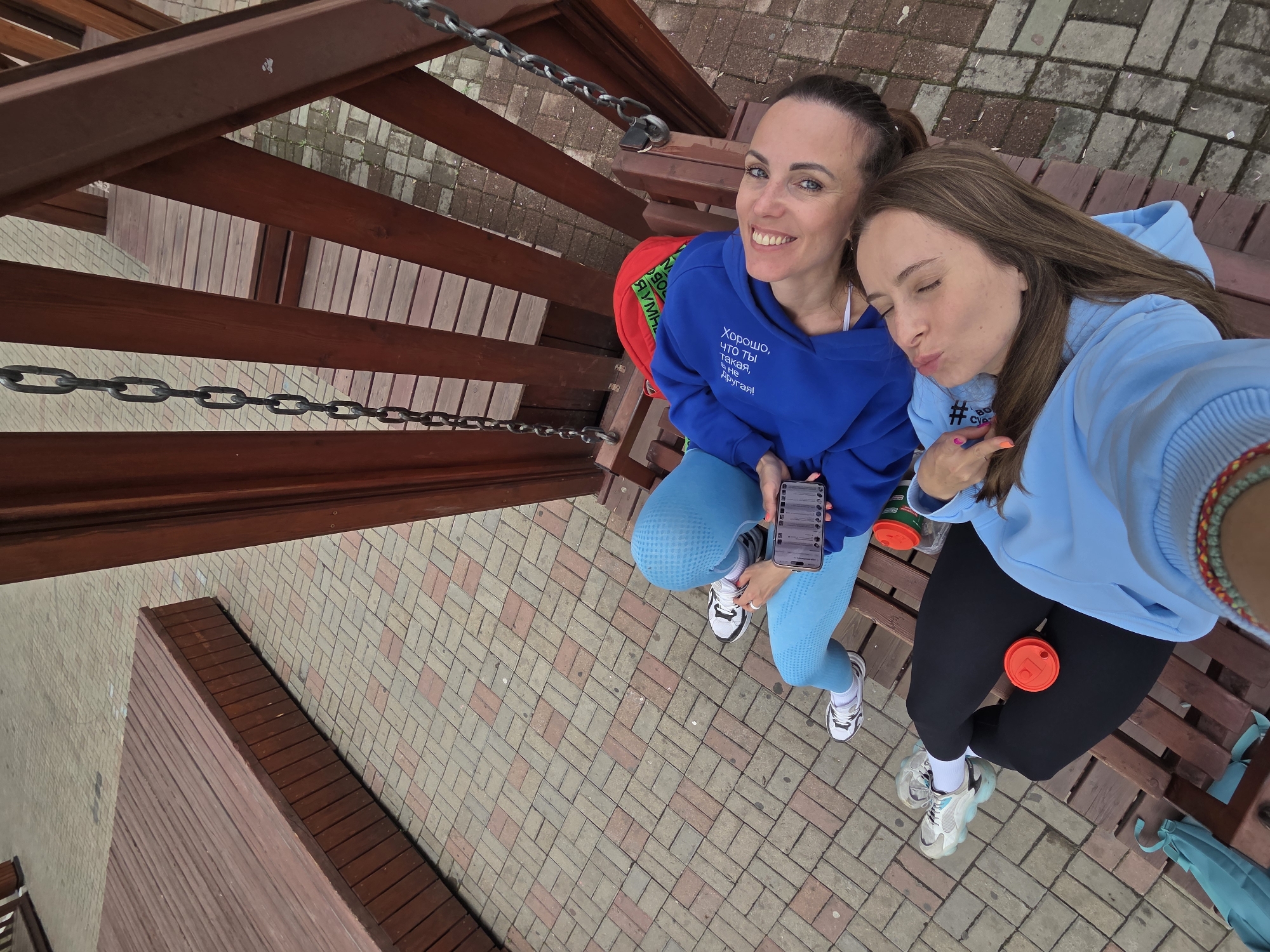 A high-angle full shot shows two women posing for a selfie on a bench on a brick-paved area, likely in Birobidzhan, Russia. The woman on the left has her hair pulled back, wears a blue hoodie with Russian text, light blue leggings, and white sneakers. She holds a phone and smiles at the camera. A red backpack sits behind her. The woman on the right wears a light blue hoodie and black leggings, also with white sneakers. She is making a kissing face and pointing with her right hand. Red cups are placed between them and on the bench. A railing with metal chains is visible above them, along with wooden benches and planks. The ground is paved with red and beige bricks.