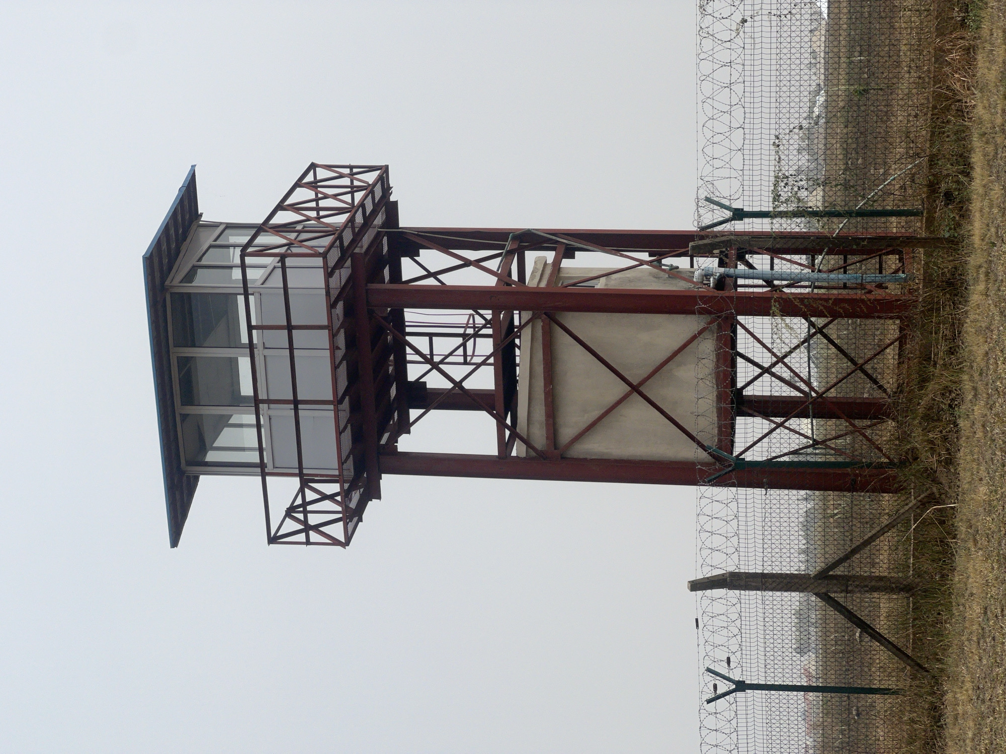 The image depicts a guard tower located next to a wire fence topped with barbed wire. The tower is constructed of red metal beams and has a glass-enclosed cabin at the top. The cabin has a blue roof and a small observation deck. The ground around the tower is dry grass and weeds. The sky is overcast. No people are visible. The location is stated to be Haifam, Nigeria.