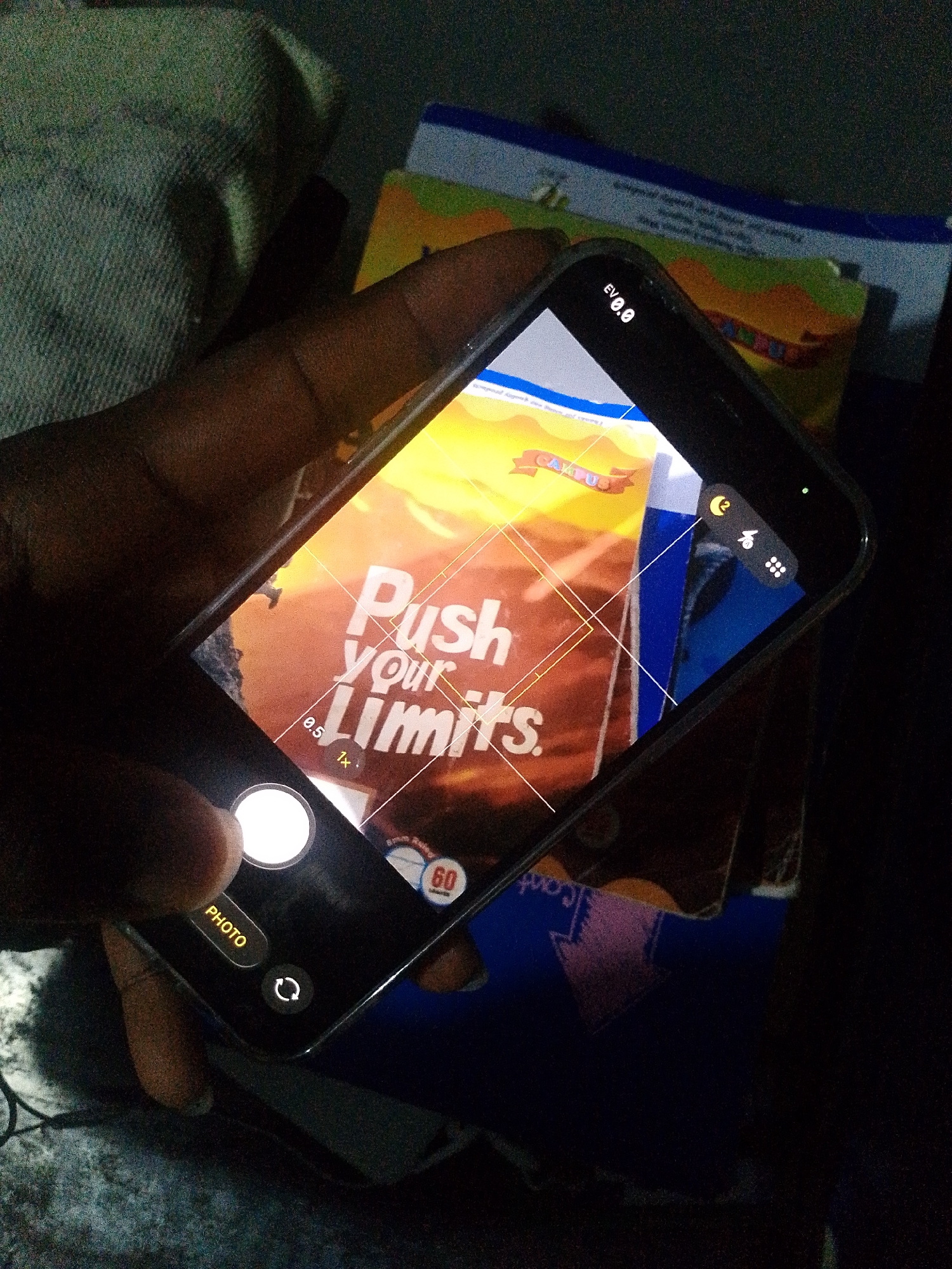 A person is holding a smartphone, taking a photo of a book with the words "Push your Limits." The book is on top of a stack of other books. The scene appears to be indoors with low lighting. The location is Ipetumodu, Nigeria.