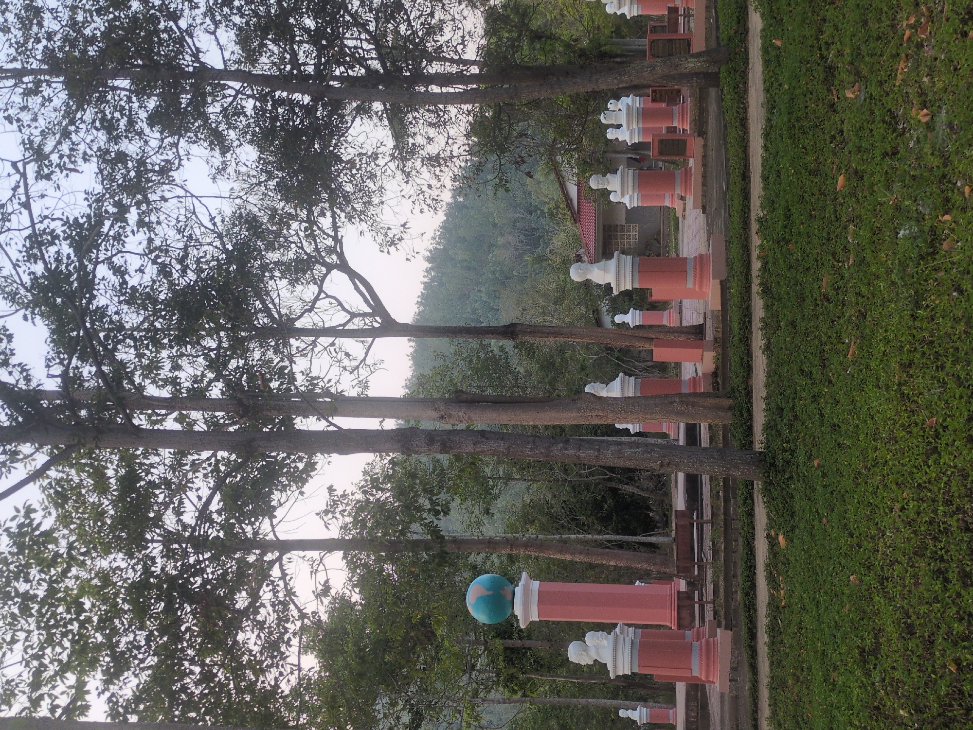 This photo depicts a gravesite in South Quy Nhon Ward, Vietnam. Several trees tower over a line of headstones, each consisting of a red pillar topped with a white decorative sculpture. One headstone features a globe with a blue and orange map. A patch of green grass borders the gravesite, contrasting with the wooded hills in the background.