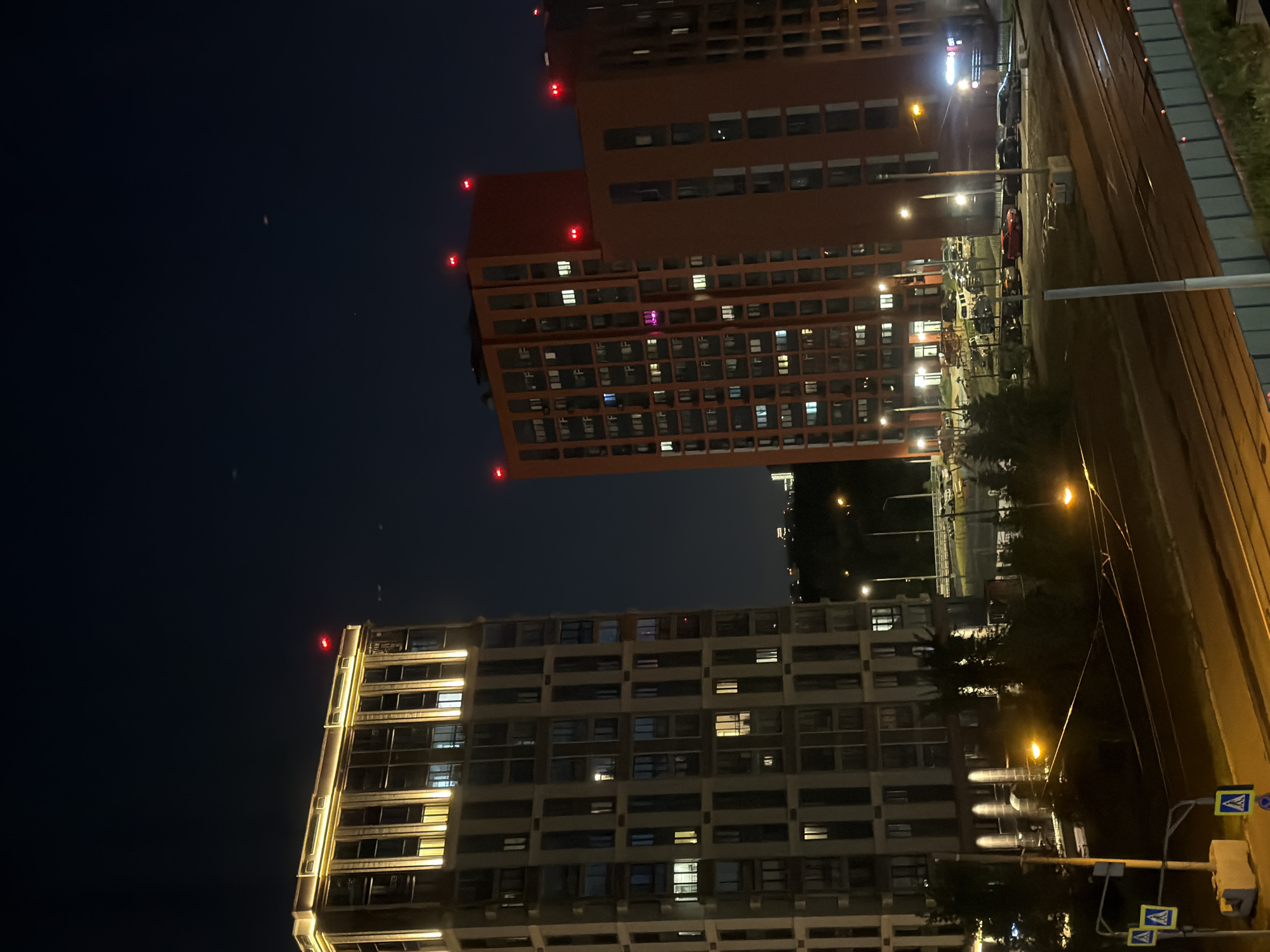 This media file depicts an urban scene at night in Moscow, Russia. The primary subjects are two large, modern residential buildings. The building in the foreground is lighter in color and illuminated with warm, yellow lights along its facade and within its many windows, suggesting occupancy. The building further back is a darker, reddish-brown color and is also lit, with some windows showing light. Both buildings have a blocky, contemporary architectural style, with many rectangular windows.
The scene is set at night, as indicated by the dark sky with faint stars and the artificial lighting from the buildings and streetlights. The presence of streetlights, power lines, and a street or road running alongside the buildings suggests an urban environment. There are also some vehicles visible parked or driving on the road, and a set of tram tracks running parallel to the road. Red warning lights are visible on the tops of the reddish-brown building, likely indicating an aviation hazard.
No people are clearly visible in the image. The overall mood is calm and nocturnal, with the lights creating a sense of urban life continuing into the night. The image's perspective is from a slightly elevated position, looking across the street towards the buildings. There is no visible text in the image.