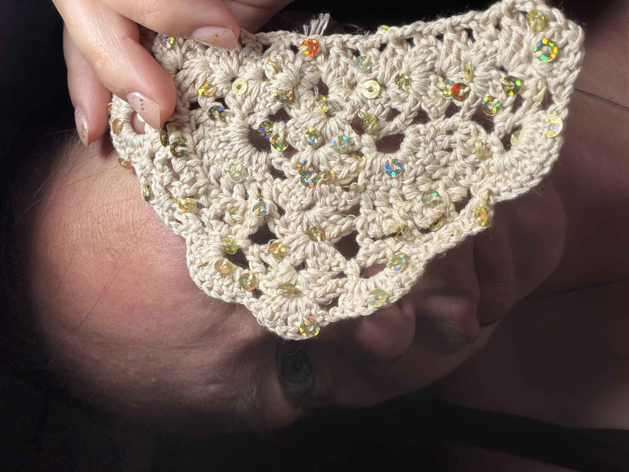 A woman's face is partially obscured by a cream-colored crocheted piece adorned with sequins, held in front of her by her hand. The crocheted piece has a scalloped edge and an openwork pattern. Her fingernails are manicured with a light color and gold flakes. The background is dark and out of focus. The scene appears to be indoors, but there is no information about the location in Chaikovsky, Russia.