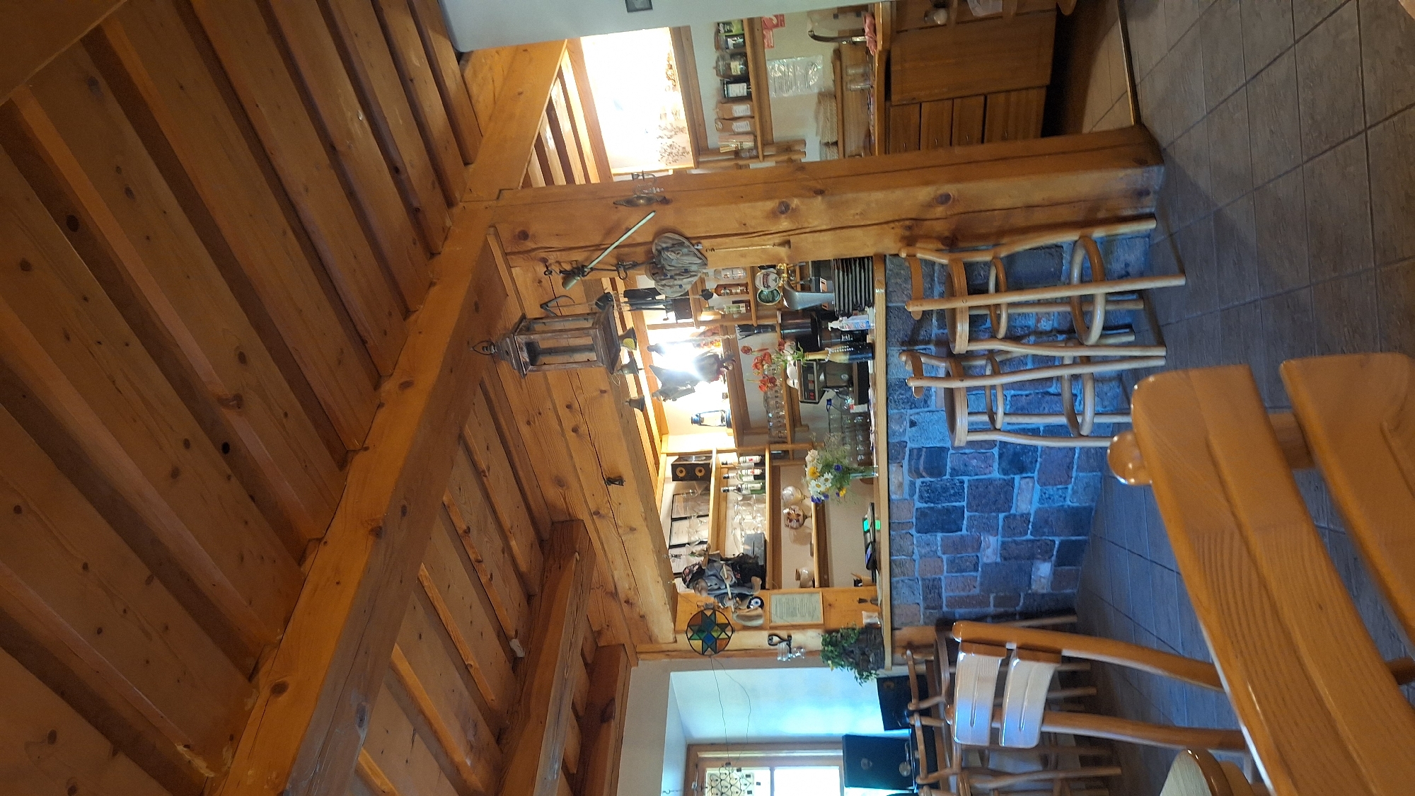 The image depicts the interior of a rustic establishment, possibly a bar or cafe, with a prominent wooden ceiling and exposed beams. A stone-clad bar counter runs across the mid-ground, behind which shelves stocked with bottles, glasses, and other items are visible. Several bar stools are placed in front of the counter. To the left, a window with decorative glass is present. In the foreground, wooden benches and chairs suggest seating for patrons. The overall ambiance is one of a traditional, cozy space. The location is identified as Lēdmane, Latvia.