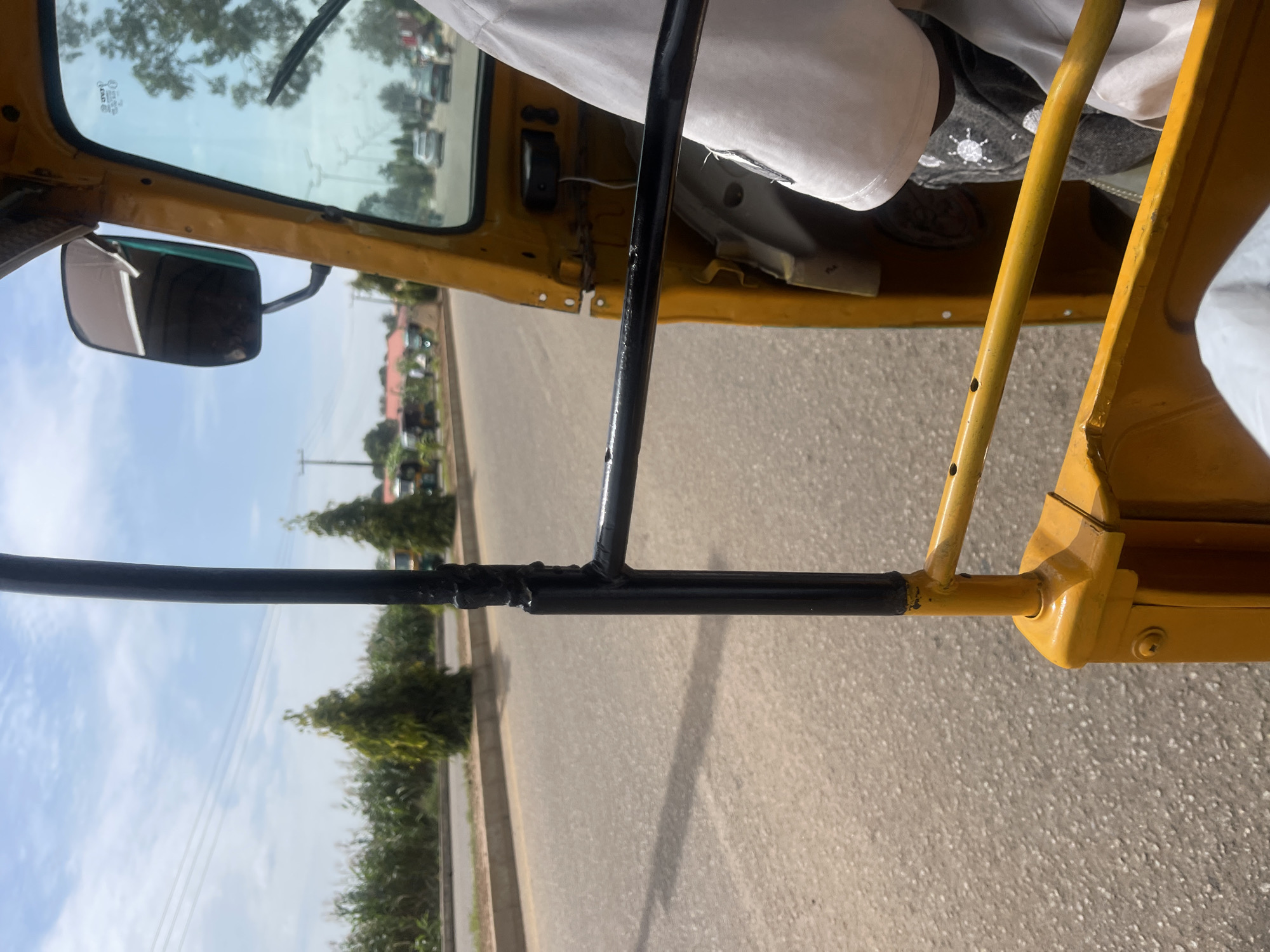 This image, captured from inside a yellow auto-rickshaw in the city of New Stadium, Nigeria, presents a view of a paved road and its surroundings. The auto-rickshaw features a black metal bar in the foreground, attached to yellow supports. A person wearing a white t-shirt and dark patterned trousers is partially visible within the vehicle, seated to the right. Through the tinted windshield, other vehicles are seen on the road, including cars and possibly other auto-rickshaws. The road is bordered by a curb and a strip of greenery, beyond which a field of tall green plants, possibly crops like maize, stretches out. Two conical evergreen trees stand beside the road, and further in the background, there are buildings and more trees, suggesting a semi-urban or rural setting. The sky is blue with scattered white clouds, and power lines are visible against the sky. The scene appears to be a snapshot of daily life and transportation in New Stadium, Nigeria.