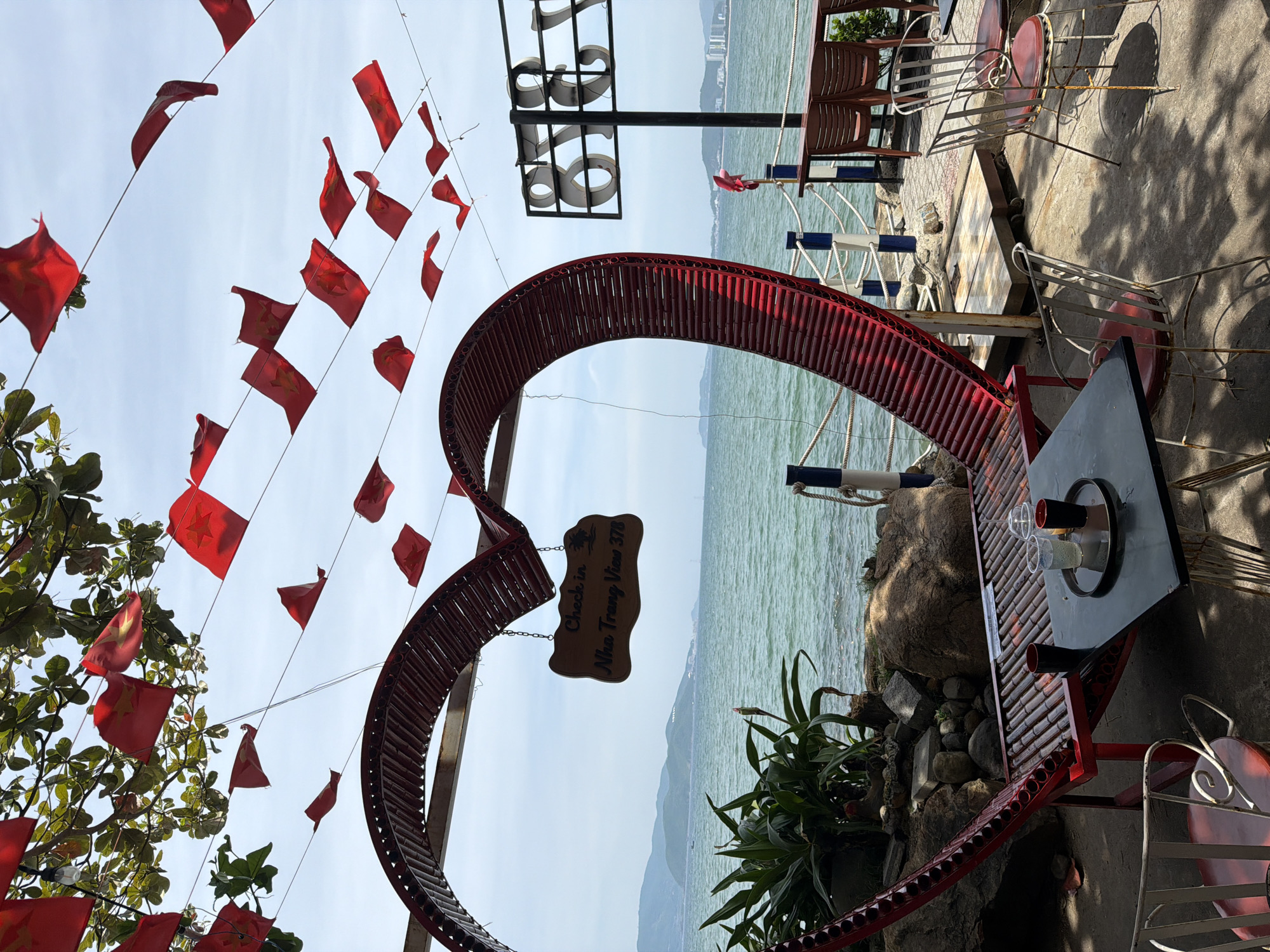 This is a ground-level, eye-level, vertical, full shot of an outdoor cafe with a view of the ocean. The cafe is decorated with numerous small red flags strung across wires overhead. A large, red, heart-shaped structure with a slatted surface serves as a bench. In front of the heart-shaped bench is a black square table with a black tray holding two cups with straws and two small black cups. Several metal chairs with red cushions surround the table. To the right of the heart structure, a sign with large, illuminated white letters reads "878". In the background, the turquoise ocean stretches out to a hazy horizon, with distant green hills and buildings. A wooden railing with white and blue posts separates the cafe area from the beach or walkway. In the foreground, on the left, there is a potted plant with large green leaves. The ground is concrete.