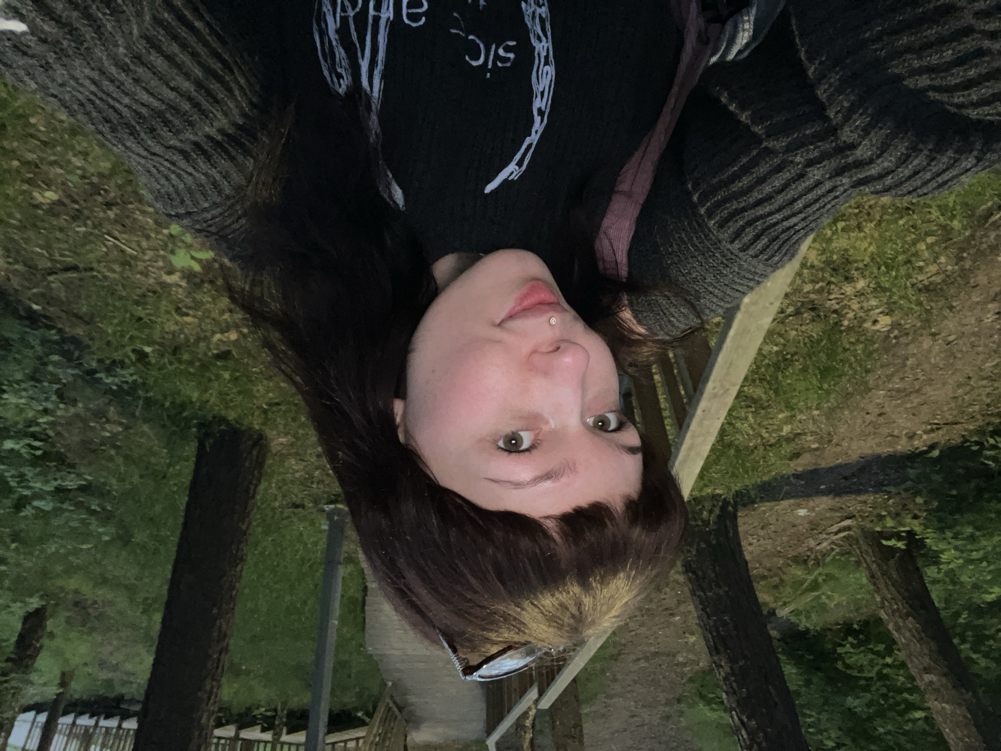 The original image is an inverted, close-up selfie of a young person outdoors. The subject has long, dark hair with bangs, light-colored eyes, and a philtrum piercing featuring a small, shiny stud. They wear a dark, possibly black, long-sleeved top with white graphic text on it; the visible inverted text includes "sic" and parts of other words. Over this, they wear a dark grey or black ribbed knitted cardigan or sweater. A pair of sunglasses rests on their head. The person looks directly at the camera with a neutral expression.
The background depicts a dimly lit natural environment, suggesting dusk or early night. Tall tree trunks and green foliage are visible, along with patches of grass and dirt. Wooden structures, resembling railings or steps, are present, indicating a constructed area within a wooded landscape. This setting is located in Chaikovsky, Russia. The entire image, including the subject and the background, is rotated 180 degrees, appearing upside down. The low light contributes to a somewhat moody atmosphere.