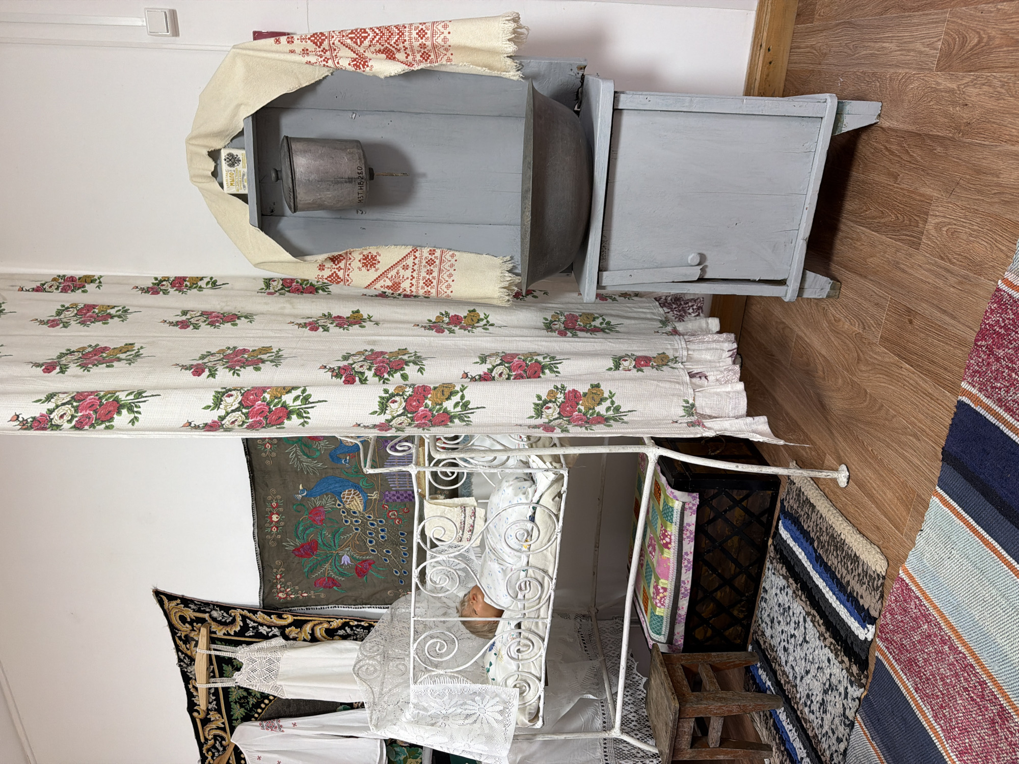 This image depicts an interior scene, possibly a museum exhibit or a preserved historical room, in Ust'-Kabyrza, Russia. The focal point is a white, ornate metal crib containing a doll dressed in white pajamas. The crib has a lace canopy. To the left of the crib, a small wooden stool sits on a woven rug. Behind the crib, a dark wooden chest is visible, topped with a colorful quilted cushion.
To the right of the crib, a tall, pale blue wooden cabinet stands against the wall. It has a small door at the bottom and a large metal basin on a shelf at the top. Above the basin, a metal pot hangs, and a small box labeled in Cyrillic, likely soap, is placed on a piece of fabric. Draped over the top of the cabinet is a cream-colored textile with red embroidered patterns, possibly a shawl or towel.
A floral-patterned curtain, featuring pink and red roses, hangs between the crib and the cabinet, creating a division in the space. On the wall behind the crib and cabinet, several decorative textiles are displayed. One is a dark tapestry with gold and black patterns, and another is a green fabric with embroidered peacocks and floral motifs. Two white garments, one with red embroidery on the sleeve and the other a white lace dress, hang on the left side of the scene.
The floor is made of wooden planks, and several woven rugs are laid out. The overall impression is of a domestic or storage setting, with elements suggesting traditional Russian or Slavic culture. The lighting appears to be artificial, illuminating the objects evenly.