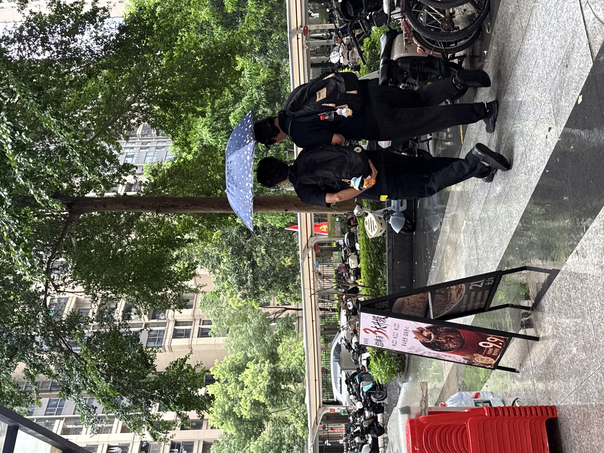 Two individuals, wearing black clothing and backpacks, stand under a blue umbrella with white polka dots. They appear to be looking down at something, possibly a menu or signage, as their heads are bowed. The scene takes place on a paved walkway, with a row of parked motorcycles and scooters visible in the background. Lush green trees and a building with numerous windows are also part of the setting, suggesting an urban environment.
A-frame signs are placed along the walkway. One sign, with predominantly red and white text and imagery, displays a close-up of a food dish and the characters "9.9". Another sign, partially visible, features text in Chinese. The presence of the parked vehicles and the overcast sky, indicated by the umbrella, suggest it might be a cloudy or rainy day. The overall impression is a brief pause or interaction on a street in a populated area, likely during daytime hours. The expressions of the individuals are not discernible.