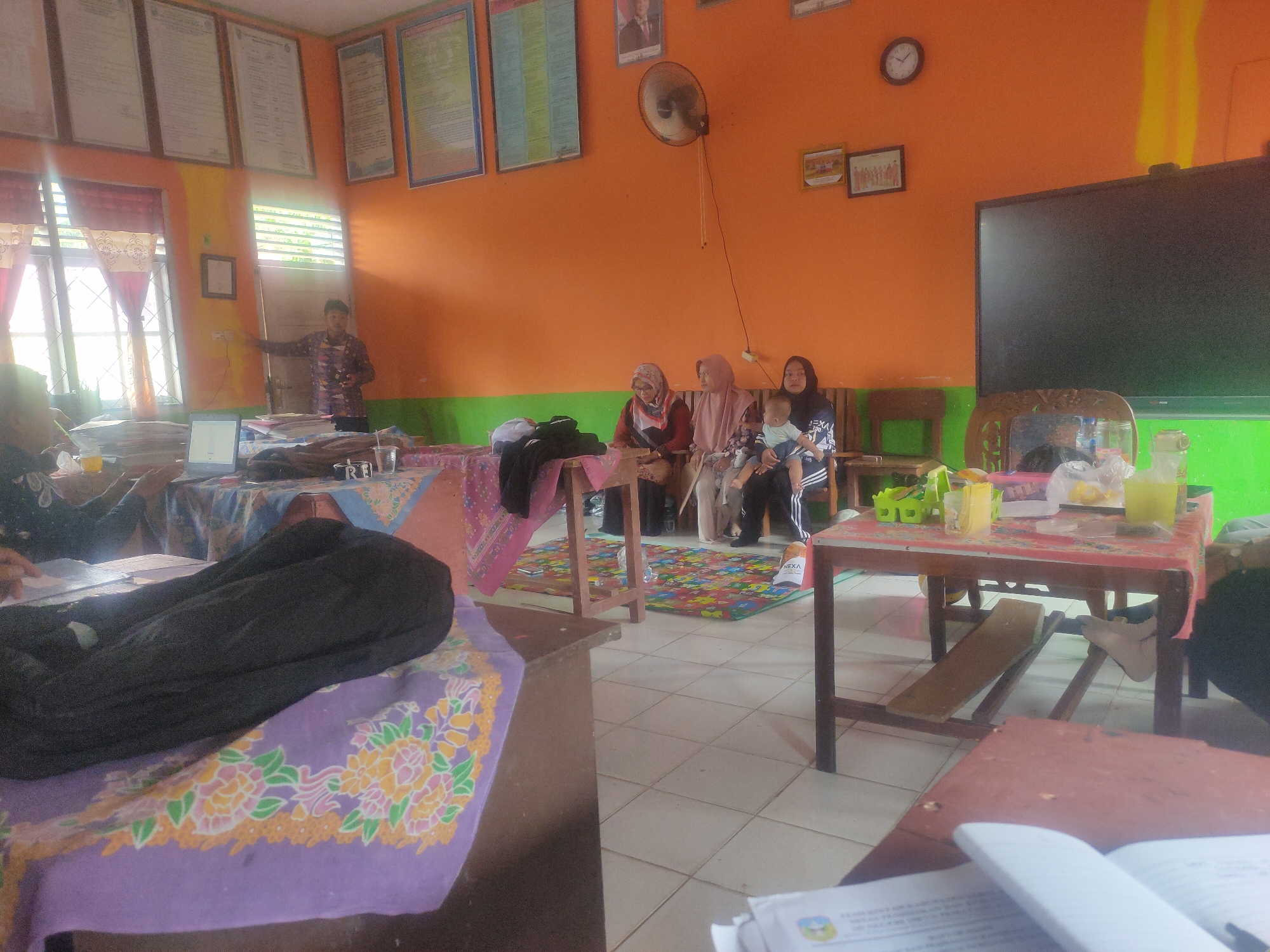 In a classroom with orange and green painted walls, likely in Pematang Kulim, Indonesia, a group of individuals are gathered. In the foreground, a person is seated with a black garment draped over their lap, resting on a table covered with a purple cloth featuring floral patterns. To the right, a small table holds various items, including plastic cups, containers, and what appears to be a bottle of yellow liquid. Behind this table, a large black screen, possibly a television or projector screen, is mounted on the wall.
Further into the room, a man in a patterned shirt stands near a window with red curtains, pointing towards something in the room. Several people, predominantly women wearing headscarves, are seated on a bench. One woman is holding a baby. Educational posters and framed pictures adorn the walls, and a circular clock hangs above them. A fan is mounted on the orange wall. The floor is tiled, and colorful mats are spread out in the area where the seated individuals are gathered. Laptops and stacks of papers are visible on tables, suggesting an educational or administrative setting.