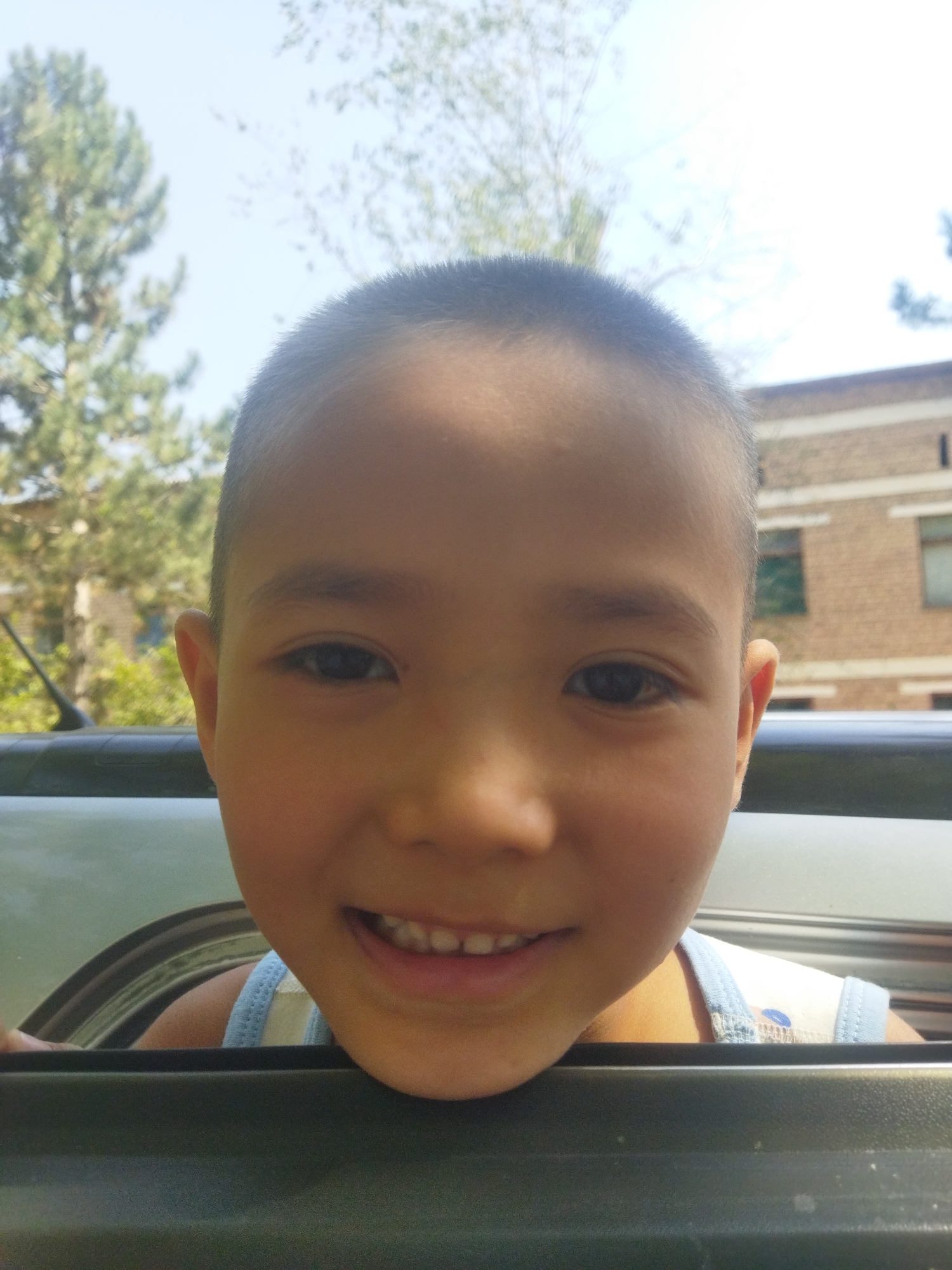 In a close-up, waist-up shot, a young child with dark hair, visible from the forehead to just below the chin, leans out of a vehicle. The child is looking directly at the camera with a wide smile, revealing teeth. The child is wearing a light blue and white striped tank top. The background is blurred, showing green foliage and a brick building. The foreground consists of the dark gray interior of a vehicle, with the child's head and shoulders positioned above it. The image appears to have been taken outdoors on a bright, sunny day, with a light blue sky visible at the top. The overall impression is a candid moment captured on a warm day.