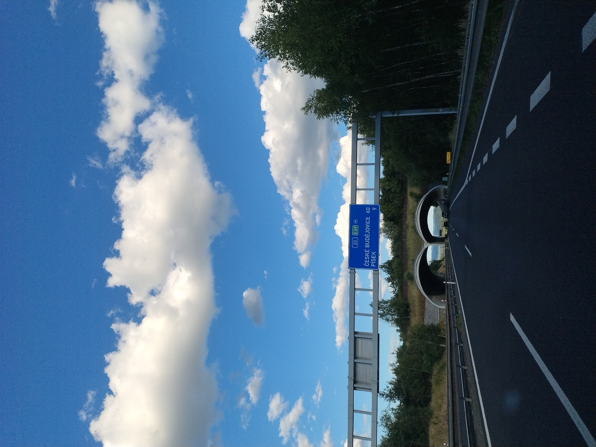 The image depicts a highway scene in Mladotice, Czechia. In the foreground, a black asphalt road with white lane markings stretches into the distance. On the right side of the road, a metal guardrail runs along the edge. Above the road, a blue highway sign indicates "ČESKÉ BUDĚJOVICE 60" and "PÍSEK 9". In the background, two arched tunnels emerge from a hillside covered in trees and grass. The sky is a vibrant blue with scattered white clouds. The overall environment is rural and appears to be during daylight hours. There are no people or specific actions captured in the image.