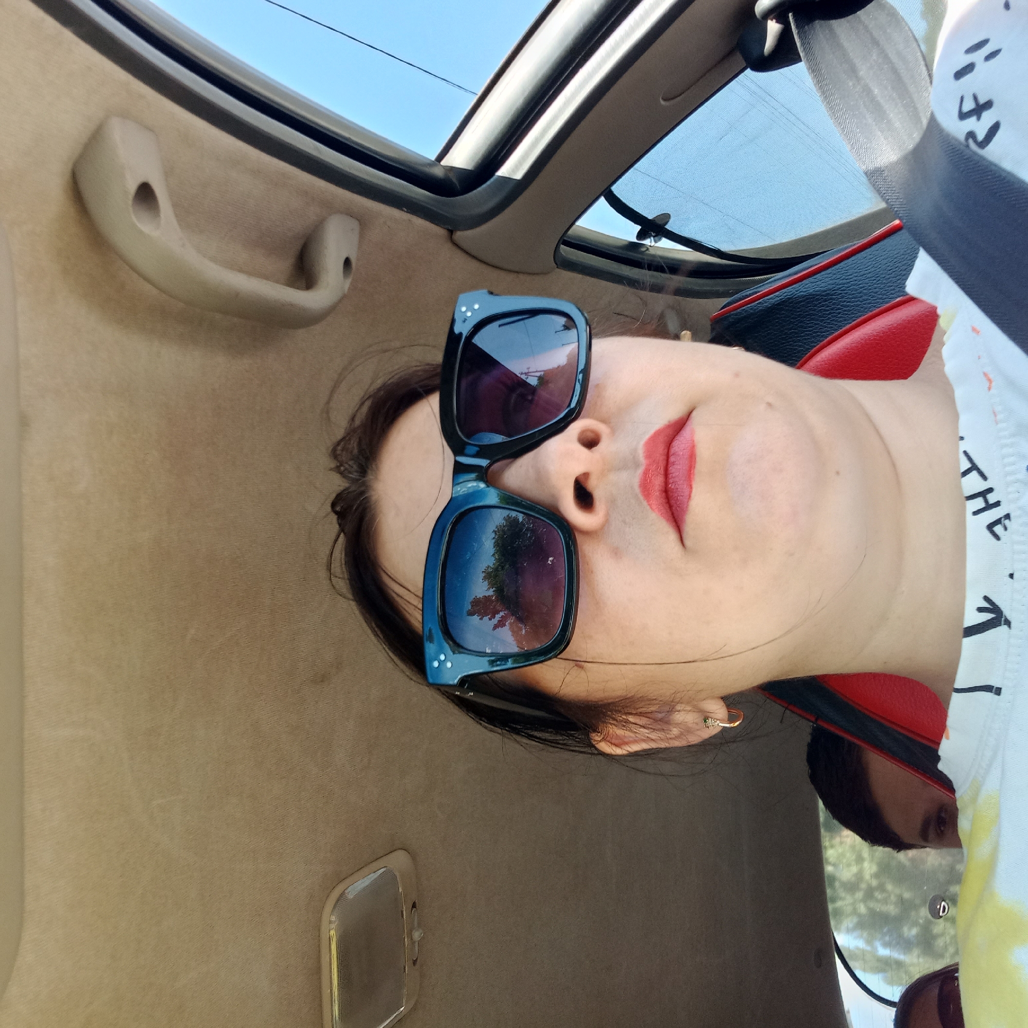 A woman wearing black-framed sunglasses and red lipstick is in the driver's seat of a car. She has dark hair and is wearing a white t-shirt with black lettering. A seatbelt is visible across her chest. In the background, a young person's head is visible between the woman and the driver's seat. The car's interior is tan, and a light fixture is visible on the ceiling. The sky outside the car window is clear and blue. The image was taken in Kropyvnytskyi, Ukraine.
