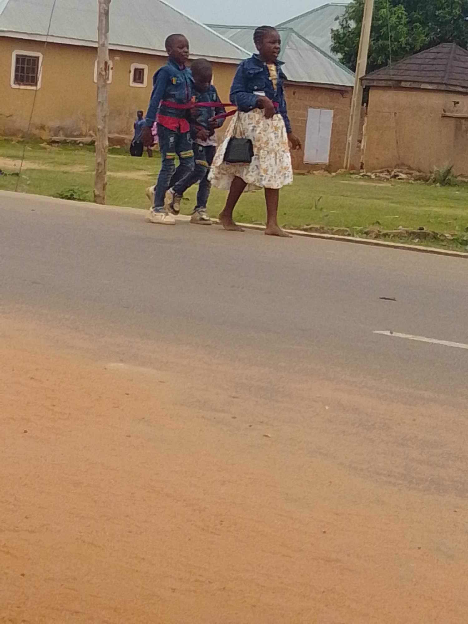A group of three young people, two boys and one girl, walk side-by-side along the right side of a paved road. The two boys are dressed in matching denim jackets and jeans, with colorful stripes on the legs of their jeans. The girl, on the right, wears a denim jacket over a floral print dress and carries a small black purse. They appear to be walking in the same direction, towards the right side of the frame. Behind them, a small, blurred figure in a blue shirt is visible near a building. The environment appears to be a rural or suburban area, with a yellow building and a brown-roofed structure in the background, surrounded by green grass and trees. The sky is overcast.