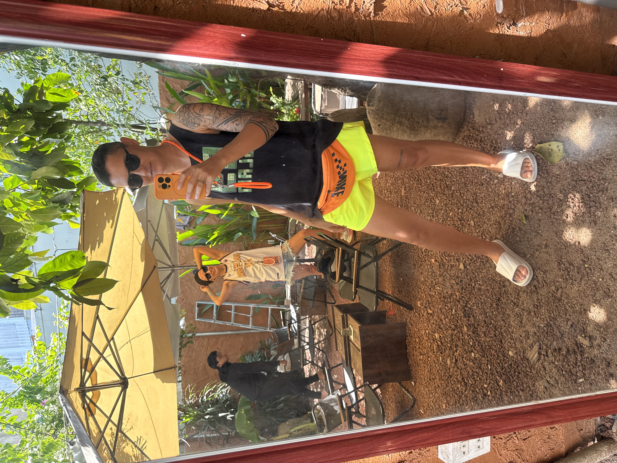 A mirror reflects a person standing outside. The person is wearing sunglasses, a black tank top, neon yellow shorts, and white slides. They are holding an orange smartphone in their right hand, taking a selfie. In the reflection, another person is visible in the background, wearing a black hoodie and pants, walking away. The environment appears to be a cafe or outdoor seating area with gravel on the ground, several chairs and tables, and greenery. An umbrella is open above the seating area. The scene is well-lit with natural sunlight.
