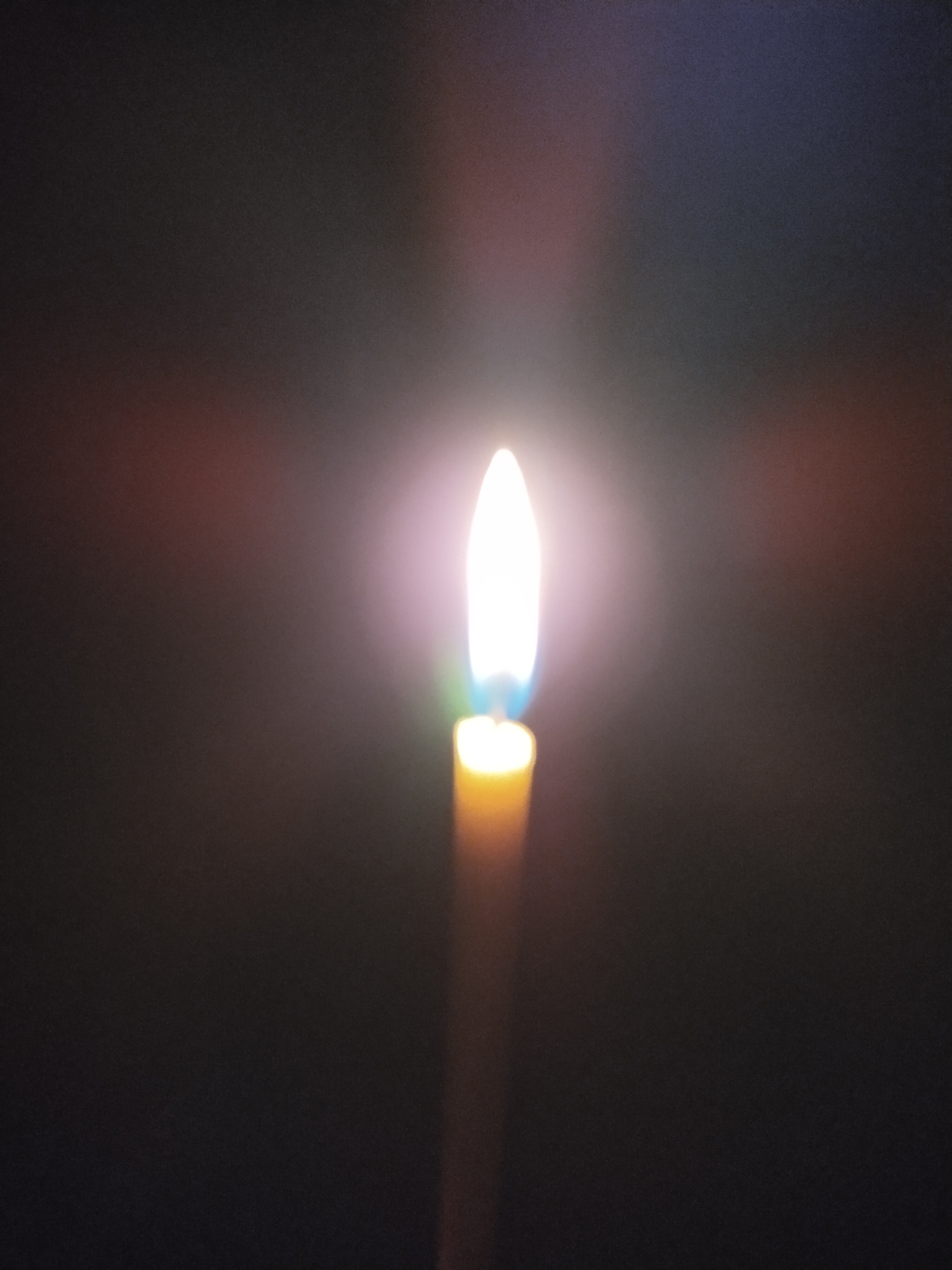 The image shows a close-up of a single candle flame burning in a dark environment. The flame is bright white at its core, with a subtle blue hue at the base and a soft pinkish glow surrounding it. The candle itself is visible as a yellow cylindrical object below the flame. The background is dark and out of focus, with some faint reddish-brown and purple light flares suggesting the presence of other light sources or reflections. The image appears to have been taken indoors, in a dimly lit room. There is no information in the image that would indicate its location in Horishni Plavni, Ukraine.
