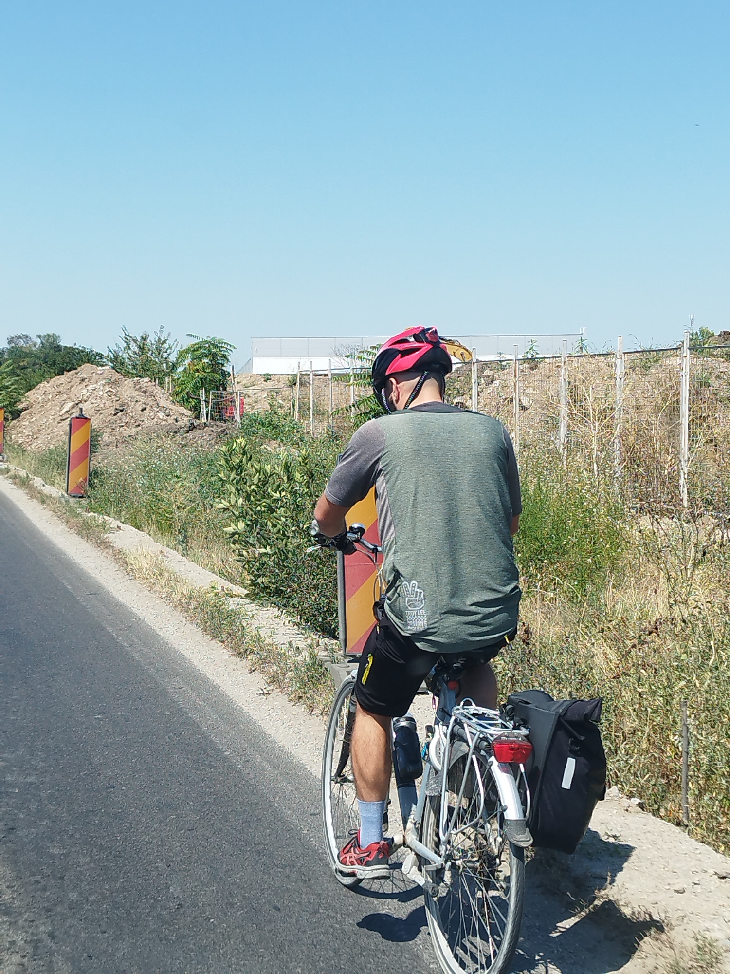 In Bragadiru, Romania, a person is cycling on a road next to a grassy embankment. The cyclist is wearing a helmet, a green t-shirt, black shorts, and red shoes. They are riding a bicycle with a rear rack and a black pannier bag. The environment shows signs of construction, with piles of dirt, construction fencing, and traffic cones visible. The sky is clear and blue, suggesting a sunny day.