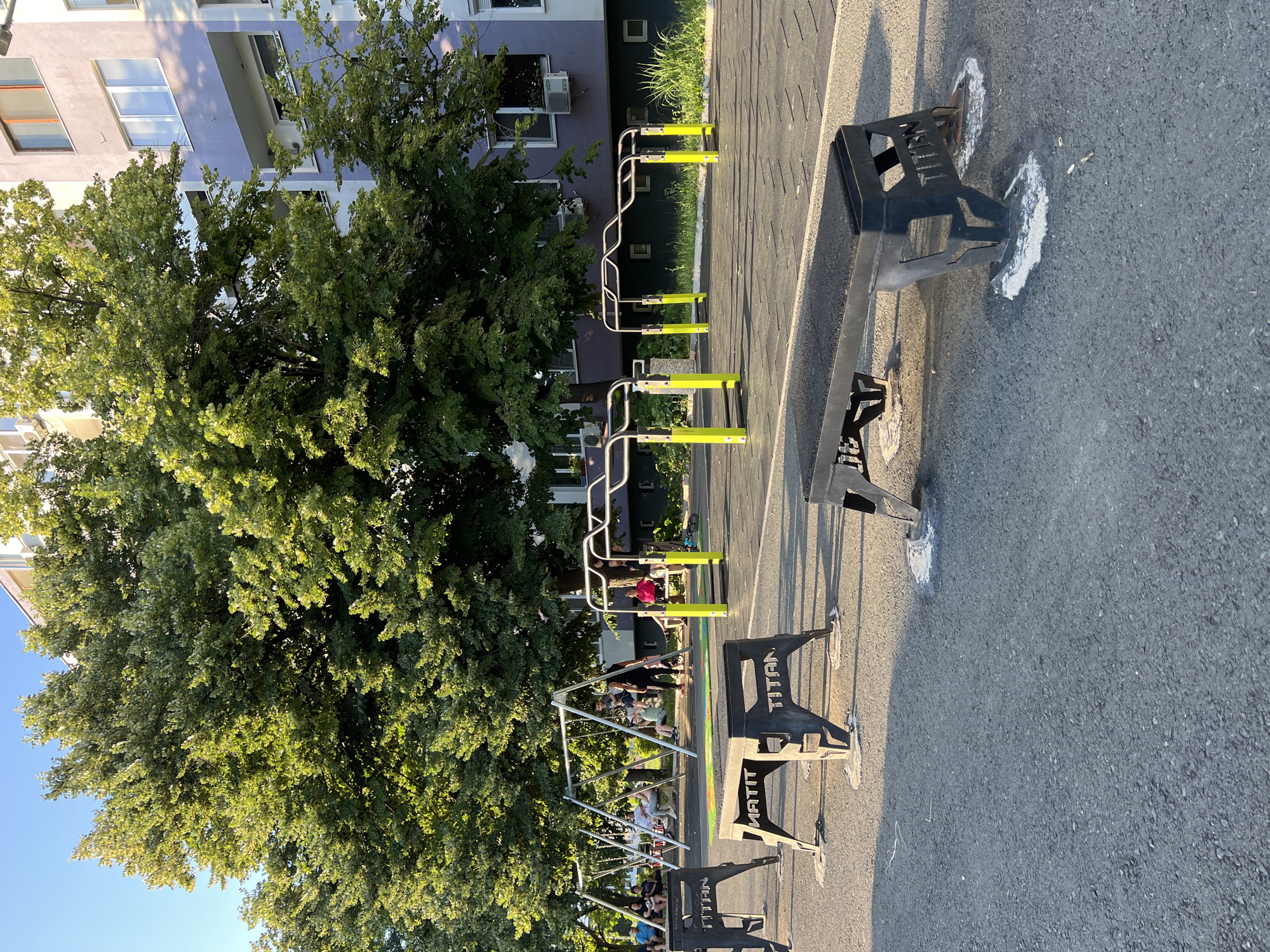 The image depicts an outdoor exercise area adjacent to a residential building. In the foreground, several black, low-profile workout benches with the brand "TITAN" are visible, secured to the asphalt ground with concrete bases. Behind these benches, a rubberized matting surface covers the exercise zone, which features parallel bars with bright yellow supports. Further back, a swing set with multiple swings is occupied by several individuals, including children and adults, engaged in recreational activity. A large, mature tree with lush green foliage dominates the middle ground, casting shadows across the scene. The background consists of a multi-story residential building with balconies and windows, suggesting an urban or suburban setting. The sky is clear and blue, indicating fair weather. This exercise area is located in the Mladost district of Burgas, Bulgaria, as indicated by the text on the benches and the architectural style of the building.