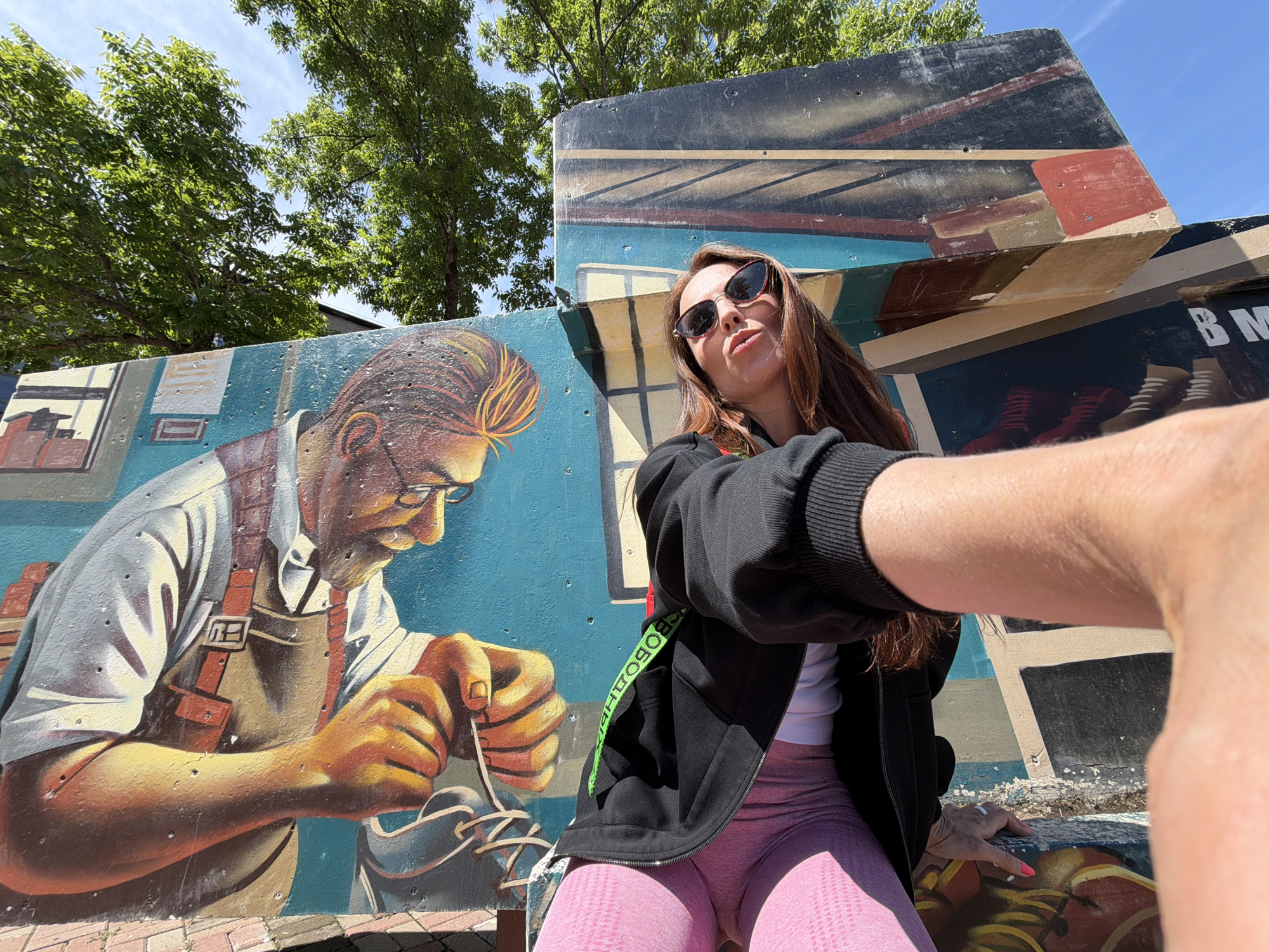 The image depicts a woman taking a selfie outdoors. She is wearing sunglasses, a black zip-up hoodie over a white top, and pink athletic leggings. She is seated in front of a mural painted on a wall. The mural features a man wearing glasses and an apron, appearing to be working on something with his hands, with tools and materials around him. The mural also has abstract geometric patterns and some text, part of which reads "BM". The scene is set against a backdrop of green trees and a blue sky. The location is identified as Birobidzhan, Russia.