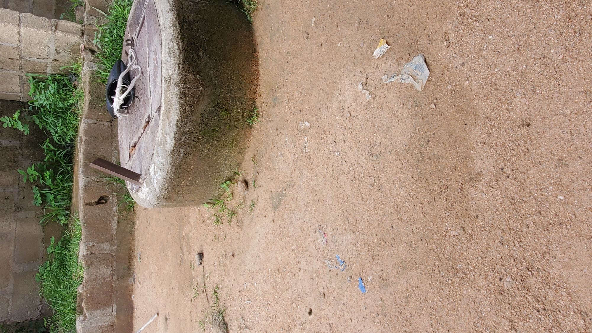 The image shows a partially covered well in an outdoor, dirt-filled area. The well is circular and constructed of concrete. Its top is covered with a metal grate, and a black object with white rope is placed on the grate. A short, metallic rod is inserted into the well's rim. The background consists of a low, concrete wall with some overgrown green plants. Scattered on the ground are small pieces of debris, including what appears to be crumpled paper and plastic. The overall scene suggests an outdoor environment, possibly in a rural or less-maintained urban area.
