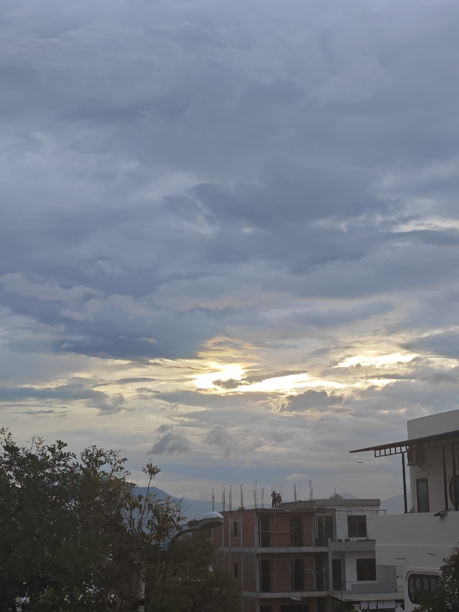 The image captures a cloudy sky over a city, likely Đà Nẵng, Vietnam, with construction sites and foliage in the foreground. The sky is dominated by heavy, gray clouds with patches of sunlight breaking through, creating a dramatic effect. Several unfinished buildings are visible, some with exposed brickwork and rebar structures, indicating ongoing construction. One building in the center features two individuals standing on its rooftop. To the right, a more completed white building with a dark canopy is present. In the lower left, dense green foliage partially obscures the view of the buildings and a streetlight. In the distance, mountains are faintly visible through the haze. A small airplane is captured in flight in the upper right portion of the frame.