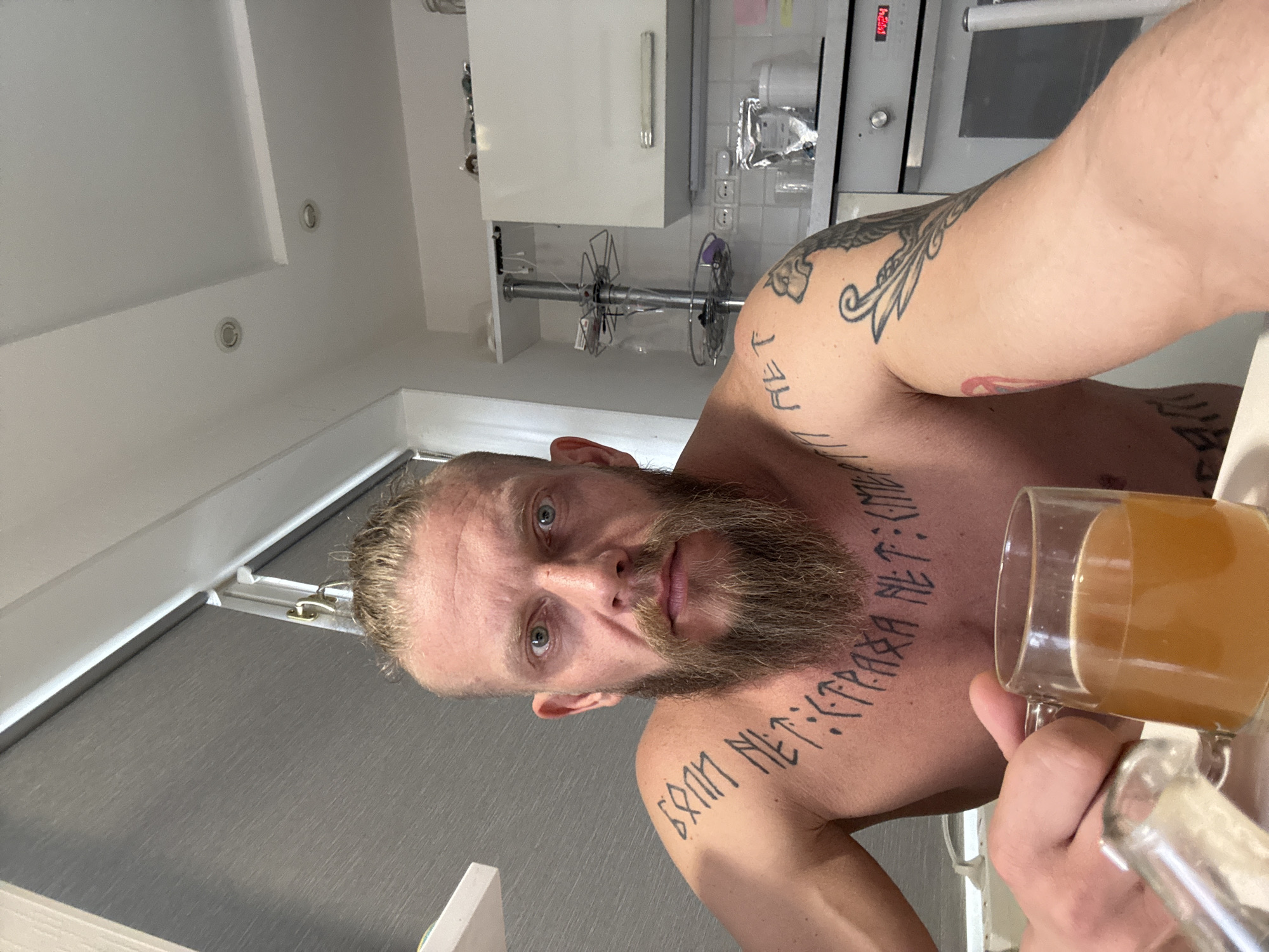 A shirtless man with a beard is holding a glass of amber liquid. He has tattoos on his chest, shoulder, and arm. He is in a kitchen with white cabinets, a metal rack, and an oven. The location is Volzhsky, Russia.