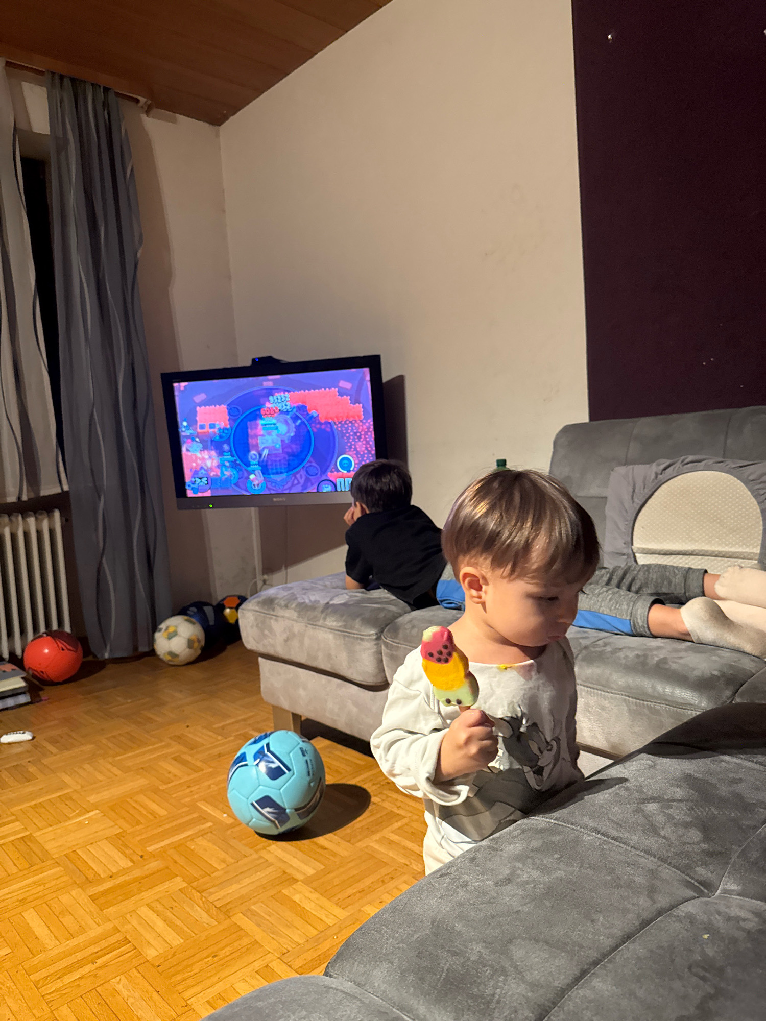 This image depicts an indoor scene, likely a living room, with a young child in the foreground holding a colorful popsicle. The child is wearing a light-colored long-sleeved shirt with a cartoon character on it. In the background, a television screen displays a video game. Another child is seated on a grey sofa, facing the television, and appears to be engaged in watching it. Scattered on the wooden floor are several soccer balls, one of which is blue and white, another is a traditional black and white, and a red ball is also visible. The room is dimly lit, suggesting it is evening. Curtains are drawn over a window, and a radiator is visible. The overall atmosphere suggests a casual, domestic setting. The presence of the soccer balls and the television indicates a common recreational space. The wooden floor and the grey sofa are key elements of the room's decor. No specific landmarks or exterior views are visible to pinpoint the exact location within Überherrn, Germany.