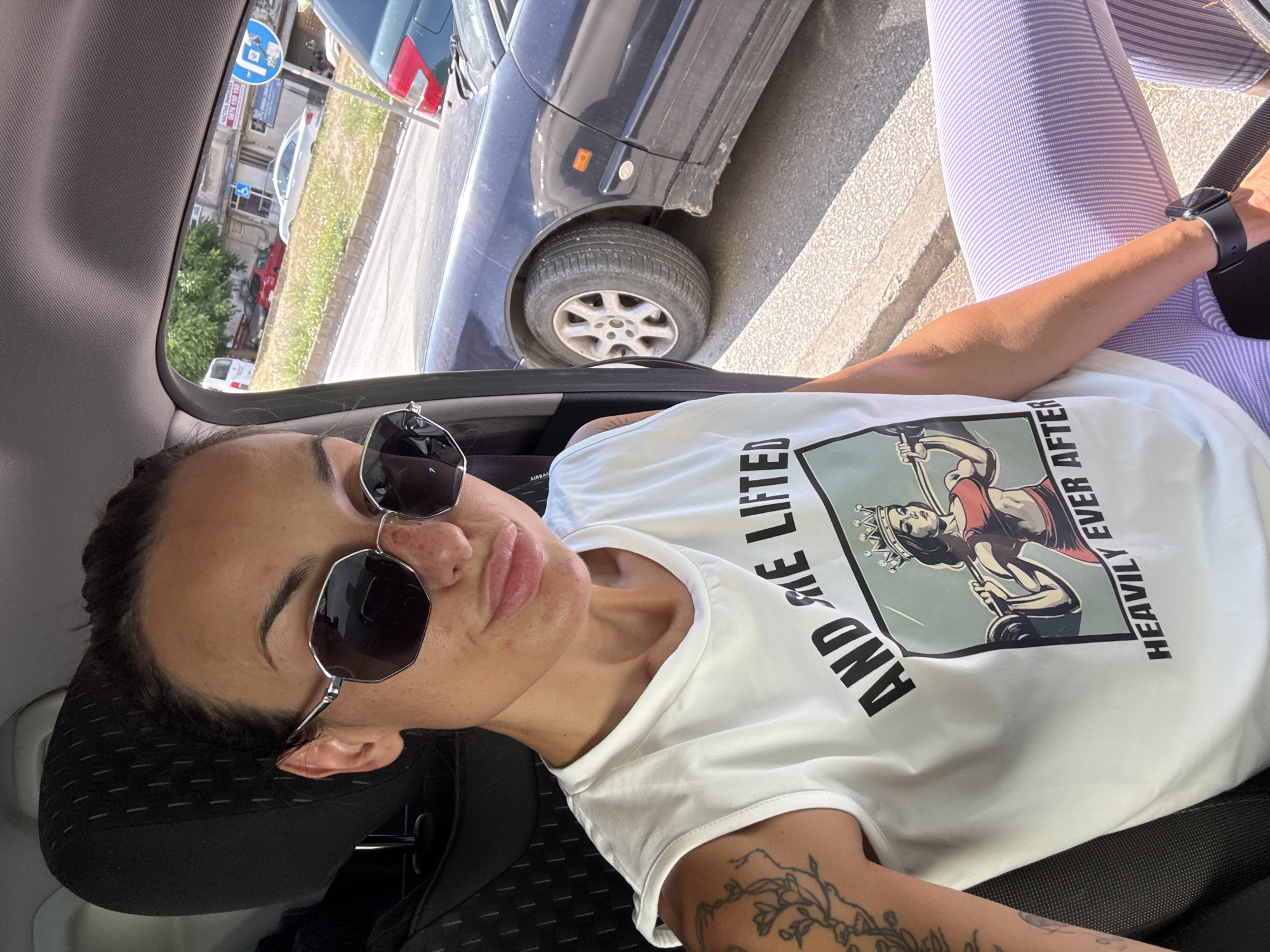 A woman sits in the driver's seat of a car, taking a selfie. She is wearing sunglasses, a white tank top with a graphic that reads "AND SHE LIFTED HEAVILY EVER AFTER" and depicts a crowned woman lifting weights, and purple striped leggings. Tattoos are visible on her left arm. The car's interior is visible, including the headrest and seat. Outside the car window, a dark blue car is parked on a street, with a sidewalk, grassy area, and buildings in the background. The scene is set in the city of Pleven, Bulgaria, as indicated by the street signs visible outside the car window. The street sign is for a parking zone.