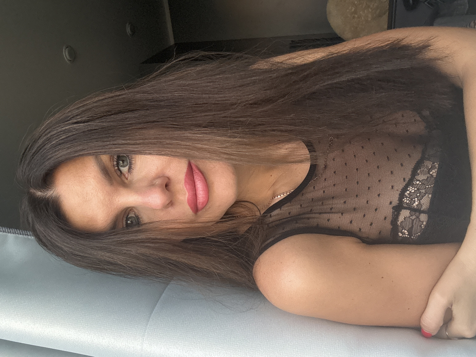 The media file is a portrait photograph of a woman. She has long, dark brown hair that falls around her face and shoulders. She is wearing a black sheer top with a polka dot pattern and lace detailing. Her makeup includes pink lipstick. Her eyes are green and she is looking towards the camera. The photograph is taken in a close-up shot, with the woman's face and upper body as the main focus. The background is dark and out of focus, with hints of a ceiling and possibly furniture. The lighting appears to be natural, possibly from a window, casting soft shadows. The orientation of the image suggests it may have been taken in a casual setting, such as a home. The woman's expression is neutral, and there are no visible objects or text in the frame. There are no clear indicators of time of day or weather conditions.