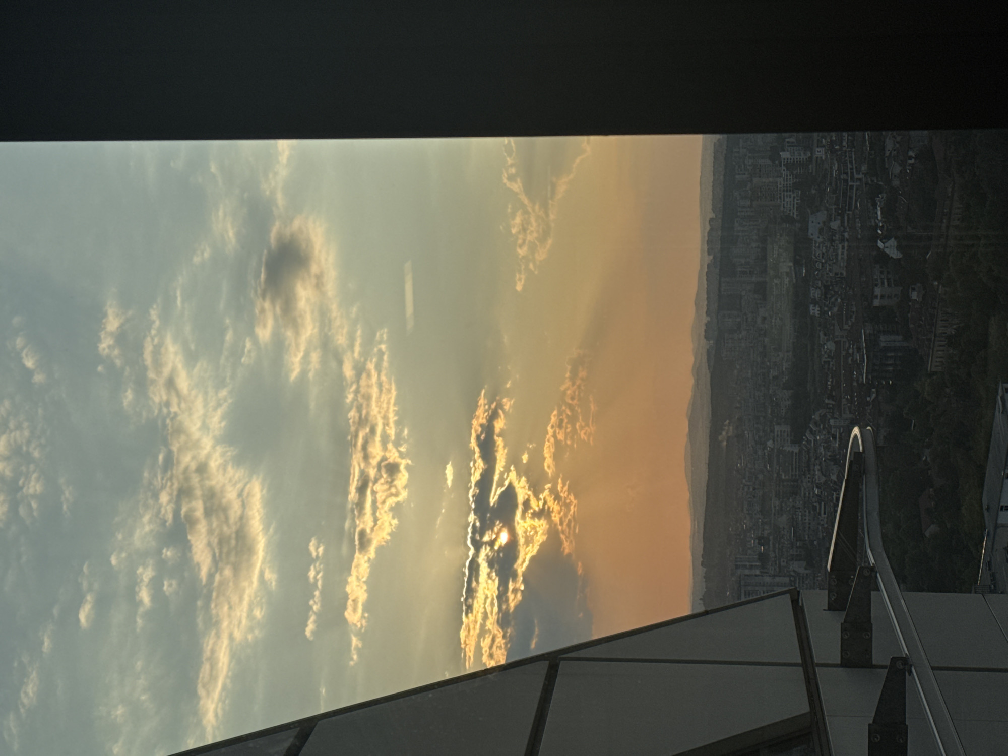 The image captures a sunset view over the city of Sofia, Bulgaria, from a high vantage point, likely a tall building. The sky is a mix of pale blue and warm orange hues, with scattered clouds illuminated by the setting sun. Rays of sunlight pierce through the clouds, creating a dramatic effect.
In the foreground, the angular structure of a modern building, likely part of the Sofia Airport Center, is visible on the left, with its metallic and glass facade. A curved metal railing or track is also present. On the right, a dark, vertical element, possibly a window frame or part of the building's exterior, creates a stark contrast and frames the view.
Below the sky, the cityscape of Sofia unfolds. Numerous buildings of varying heights are densely packed, interspersed with areas of greenery. In the distance, rolling hills or mountains form the horizon. The overall impression is of an urban environment bathed in the soft, warm light of dusk. No people are visible in the image.