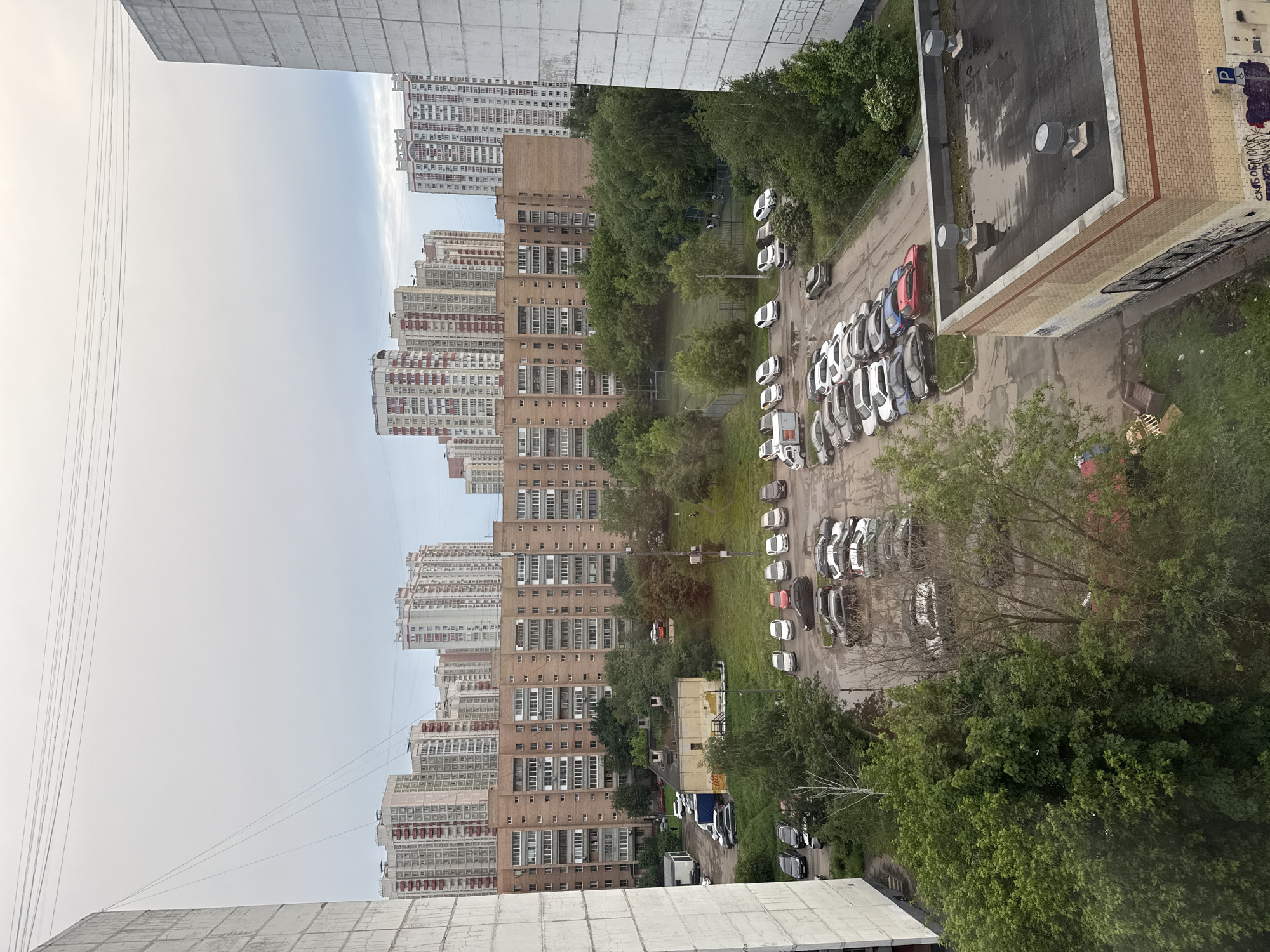 The image shows a view from a high vantage point, likely an apartment building, overlooking an urban area. Several tall apartment buildings with repetitive patterns of windows and balconies dominate the skyline in the distance. Between the buildings, there are patches of green indicating parks or tree-lined streets. A parking lot is visible below, filled with numerous cars of varying colors. There is a building with a flat roof and a brick facade on the right side. The sky is overcast. In the foreground, the edge of another building is visible, providing a sense of the viewer's location.