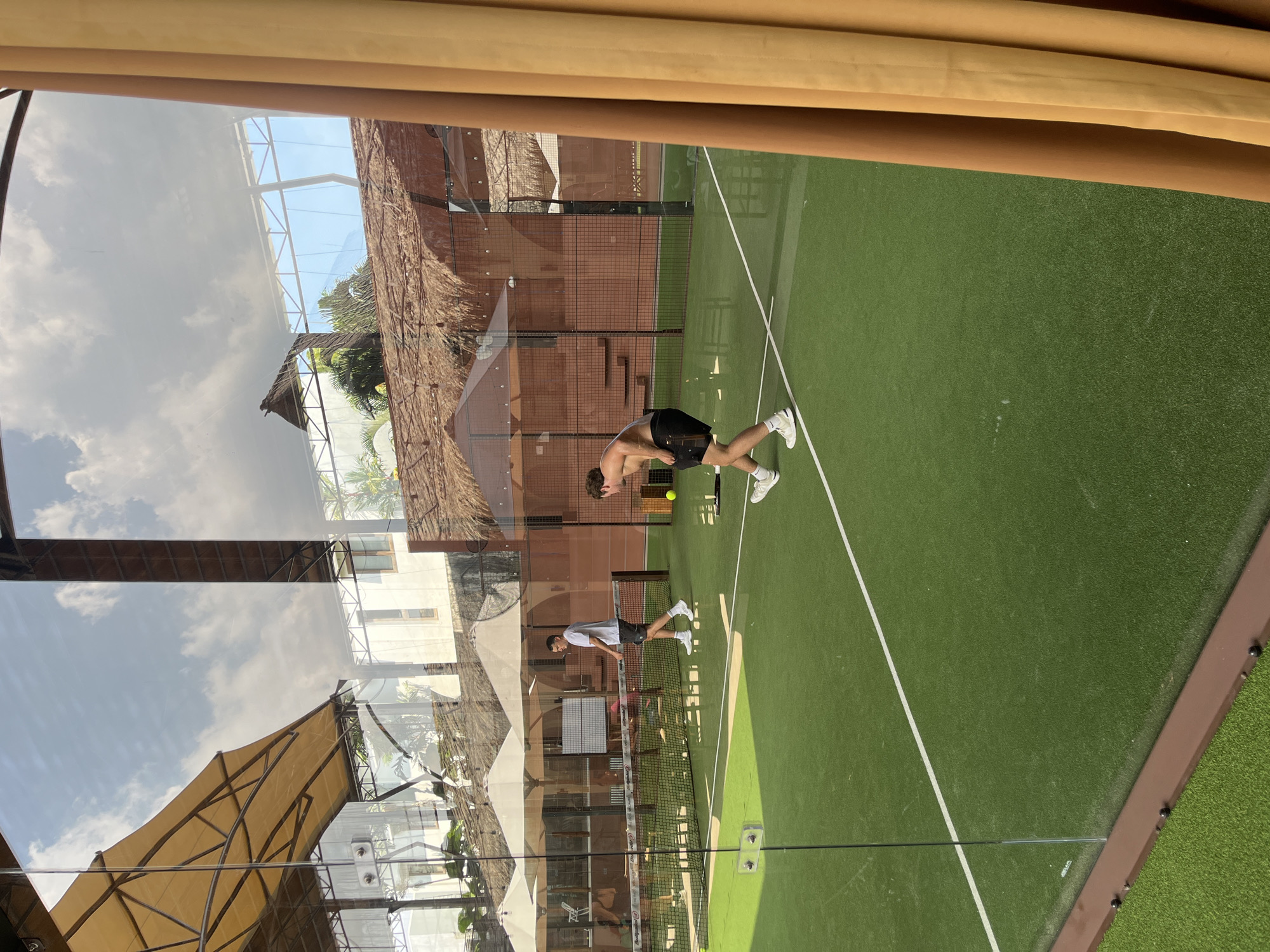 The image captures an outdoor scene, likely a sports court, in what appears to be North Kuta, Indonesia, given the tropical architecture and vegetation visible. A shirtless man wearing dark shorts and white shoes is shown in motion, presumably hitting a tennis ball with a racquet. Another individual wearing a white t-shirt and dark shorts, also with a racquet, is seen in the background. The court is green with white lines, enclosed by brown fencing. The backdrop includes structures with thatched roofs and tropical plants, reflecting the Indonesian location. The upper portion of the image shows a reflection of sky and clouds.