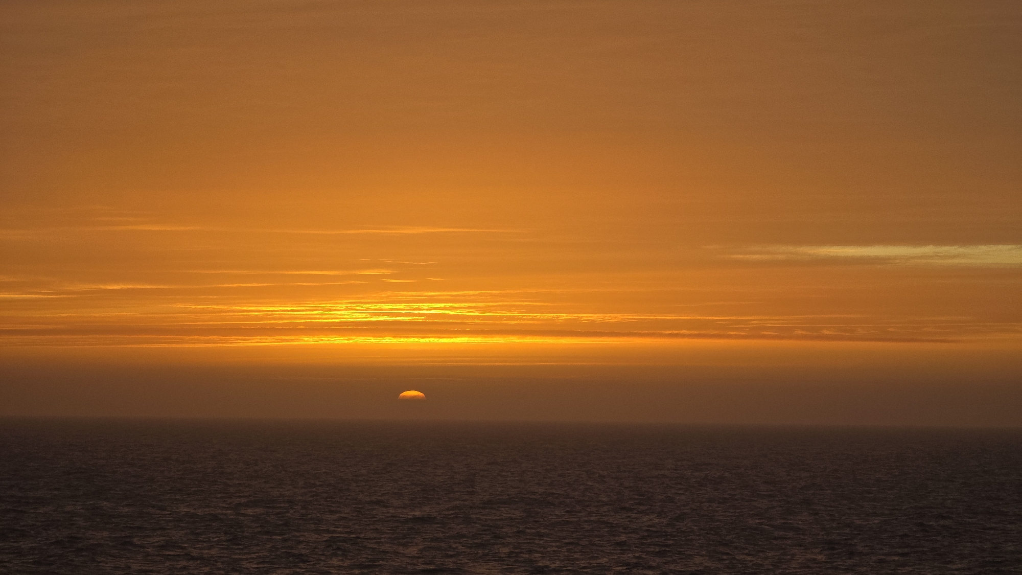 The image depicts a sunset over the ocean. The sky is a gradient of orange, from a bright, almost yellow hue near the horizon to a darker, muted orange at the top of the frame. Thin, elongated clouds stretch horizontally across the sky. The sun is a small, partially submerged orange orb on the horizon. The ocean water is dark, with subtle ripples and textures suggesting movement. The location is Unknown, Unknown.