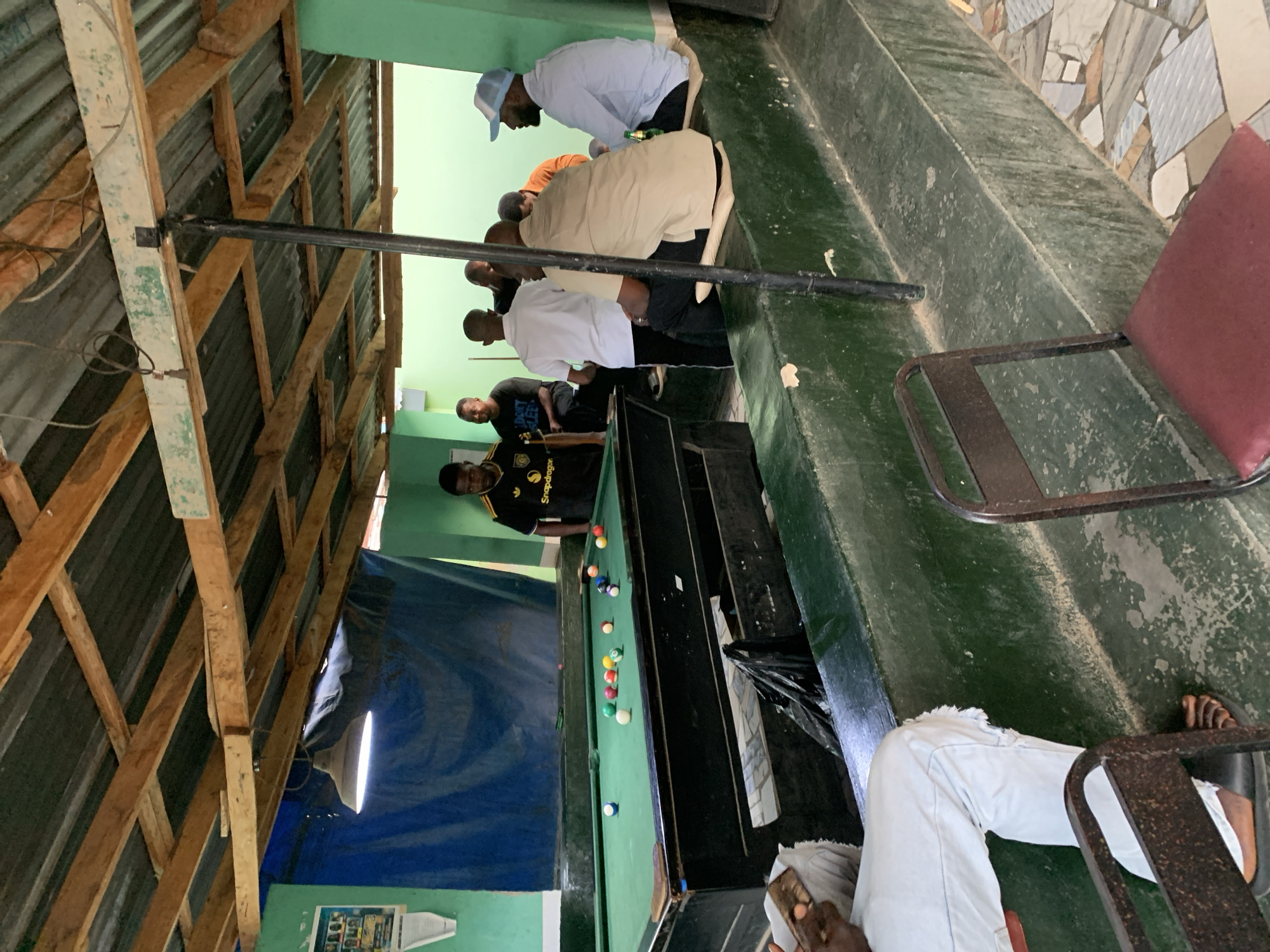 The image shows the interior of a pool hall at New Stadium, Nigeria. A pool table with several balls scattered across its green felt is the central feature. A man in a black shirt with yellow detailing appears to be playing. A group of men are watching the game from the side, some kneeling. The ceiling is made of corrugated metal supported by a wooden frame, and a light fixture illuminates the pool table. A blue sheet hangs in the background. The walls are green. A tiled area is visible near the back of the room, and chairs are placed around the perimeter.