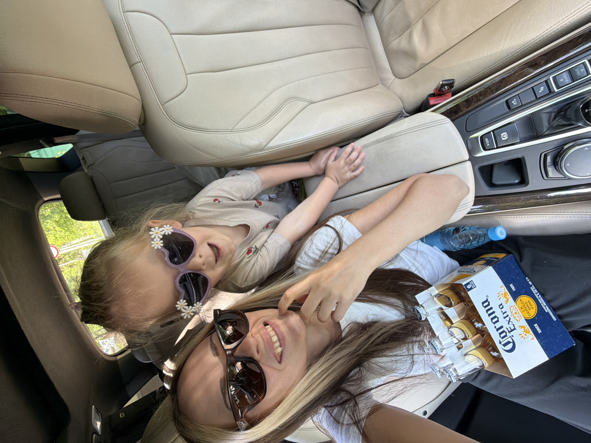 The image shows a young girl with flower-adorned heart-shaped sunglasses and a woman with sunglasses, both lying on the seats of a car. Beside the woman is a blue and white box of Corona Extra beer and a bottle of water. The scene is inside a vehicle, evidenced by the beige leather seats and car console visible in the background. The location is not identifiable as Udelnaya, Russia from the visual information provided.